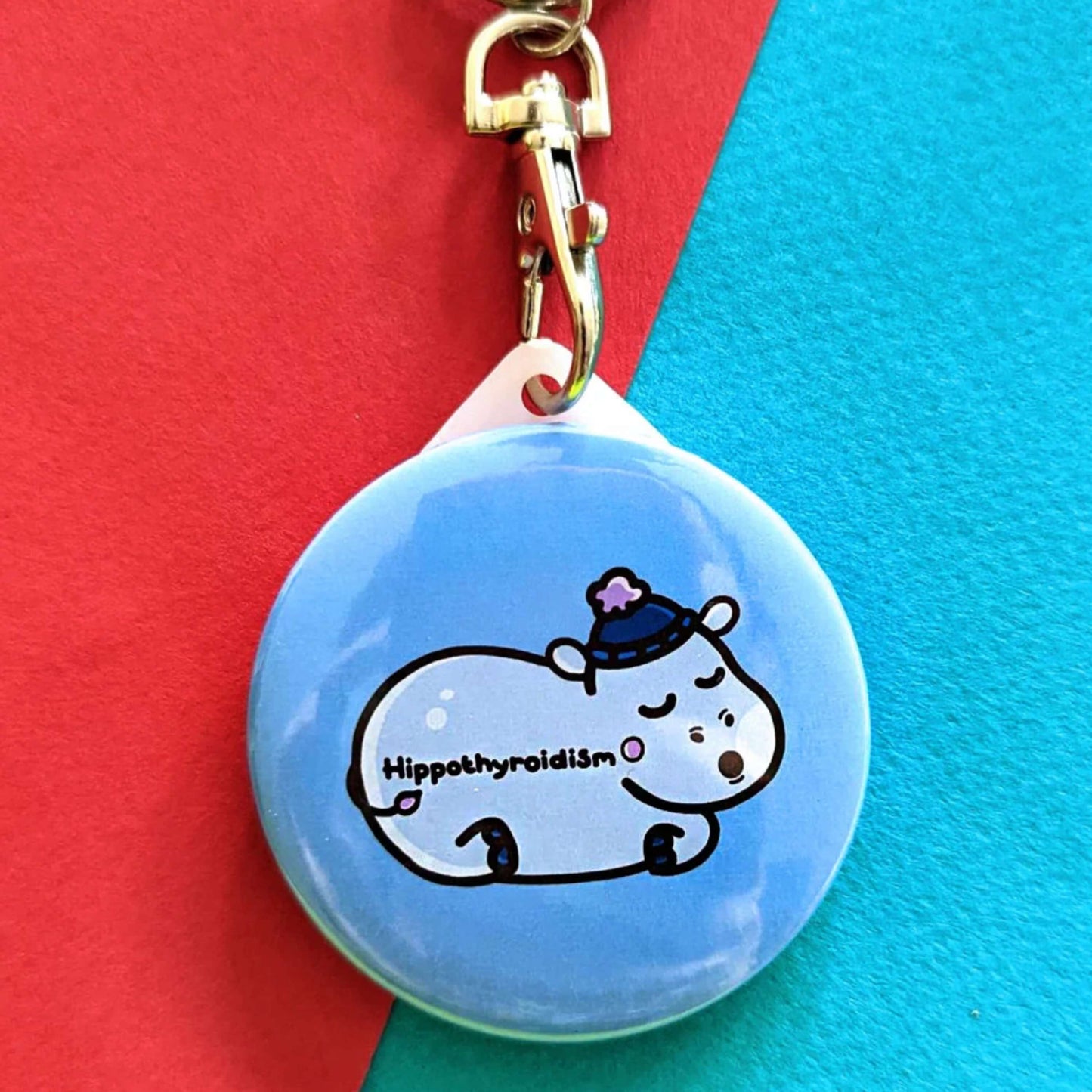 Hippothyroidism Keyring - Hypothyroidism on a red and blue background. The circular blue key ring has an illustration of a sleeping hippo wearing a blue wooly hat with Hippothyroidism written across its body. The hand drawn design is made to raise awareness for Hypothyroidism.