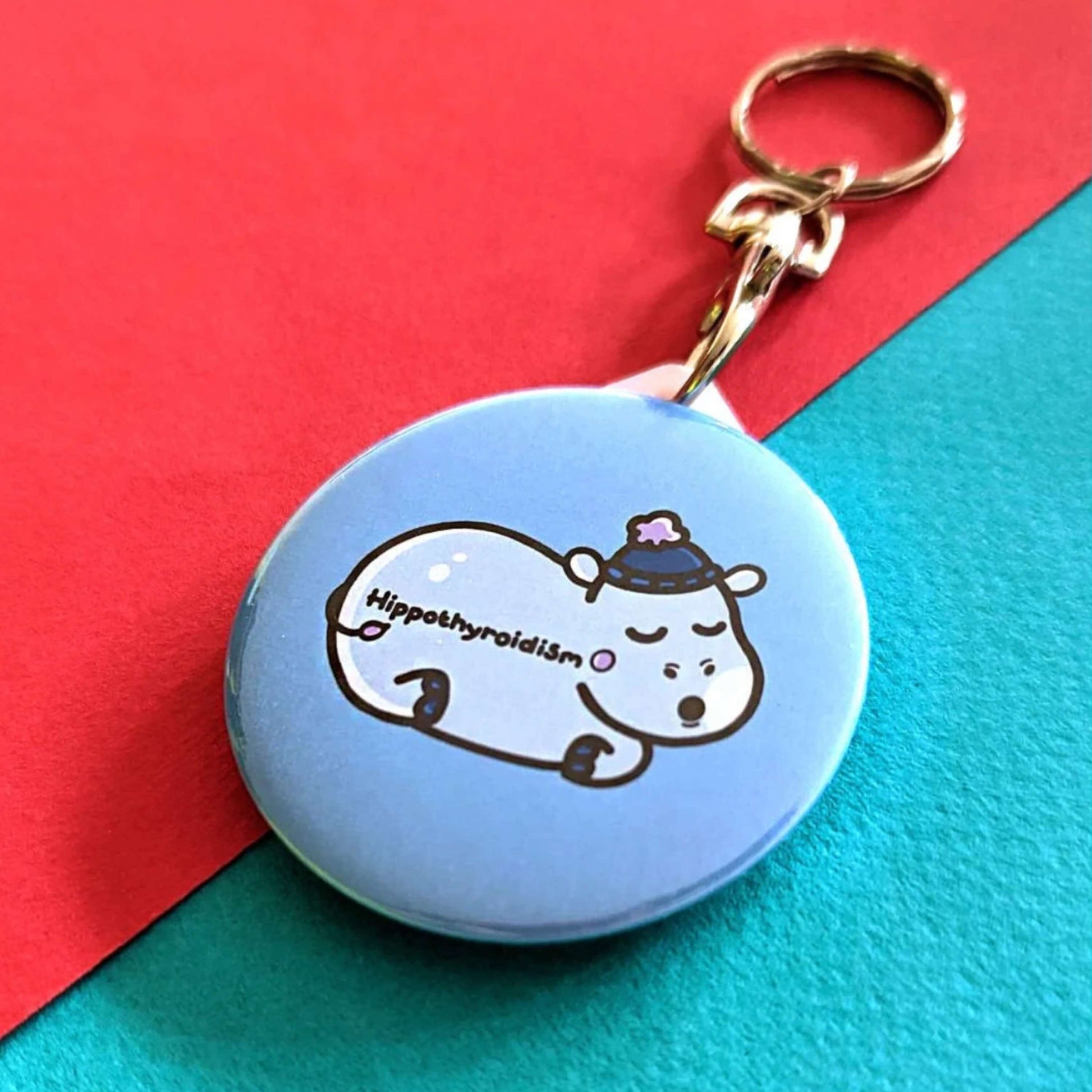 Hippothyroidism Keyring - Hypothyroidism on a red and blue background. The circular blue key ring has an illustration of a sleeping hippo wearing a blue wooly hat with Hippothyroidism written across its body. The hand drawn design is made to raise awareness for Hypothyroidism.