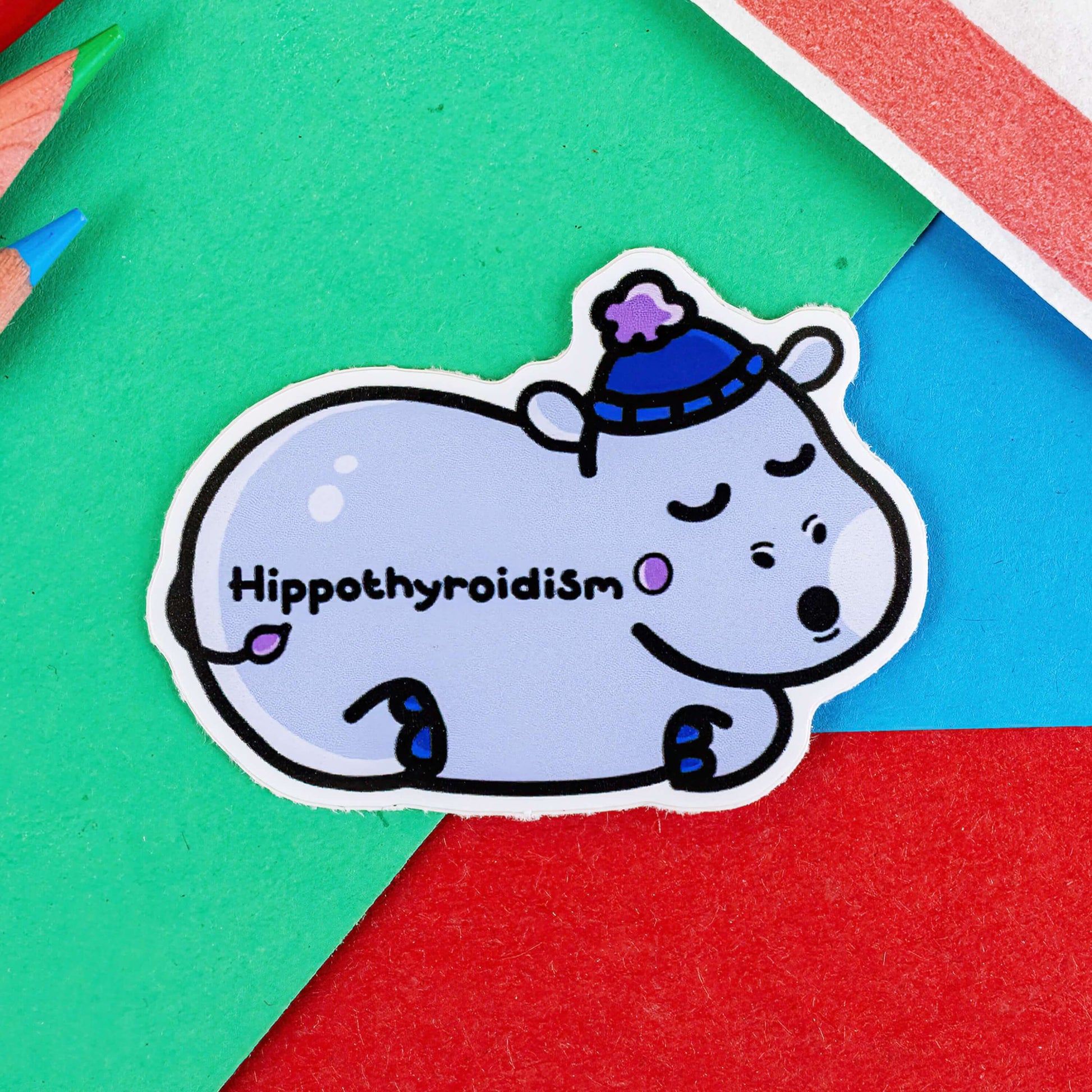 Hippothyroidism Hippo Sticker - Hypothyroidism on a red, blue and green background with colouring pencils and red stripe candy bag. A sleeping pastel blue Hippopotamus wearing a blue bobble hat shape sticker with black text across its body reading 'hippothyroidism'.  The hand drawn design is raising awareness for hypothyroidism.