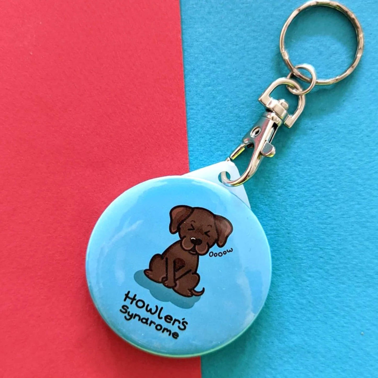 Howlers Syndrome Keyring - Fowlers Syndrome shown on a red and blue background. The circular blue keyring has an illustration of a brown dog with a pained expression and paws over its bladder. 'Howler's Syndrome' is written underneath the dog in black text. The hand drawn design is made to raise awareness for Fowlers Syndrome.