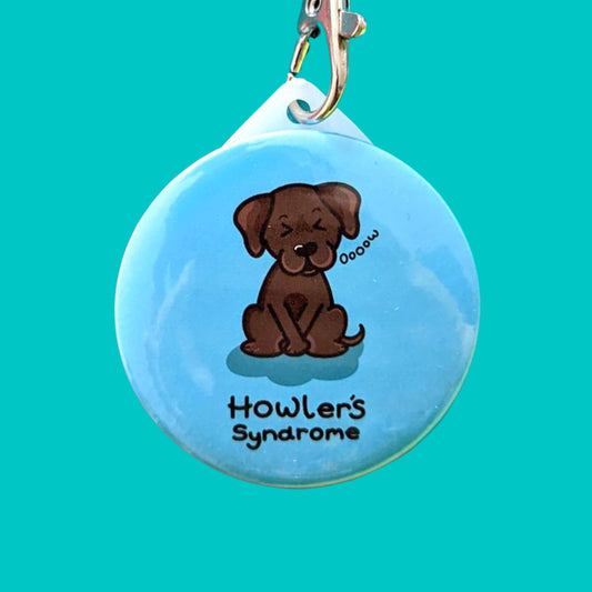 Howlers Syndrome Keyring - Fowlers Syndrome shown on a blue background. The circular blue keyring has an illustration of a brown dog with a pained expression and paws over its bladder. 'Howler's Syndrome' is written underneath the dog in black text. The hand drawn design is made to raise awareness for Fowlers Syndrome.