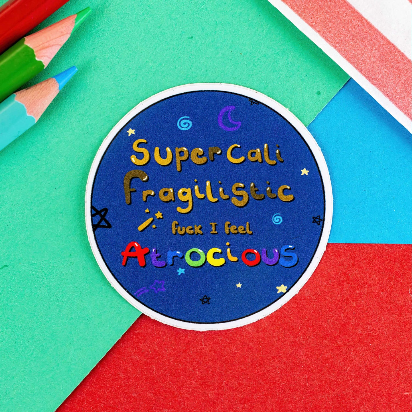 I Feel Atrocious Sticker - Blue circle sticker with the words supercali fragilistic fuck I feel atrocious' on. It is on a green, red and blue background with pencils in in the corner of the shot. Sticker designed to raise awareness for invisible illnesses.