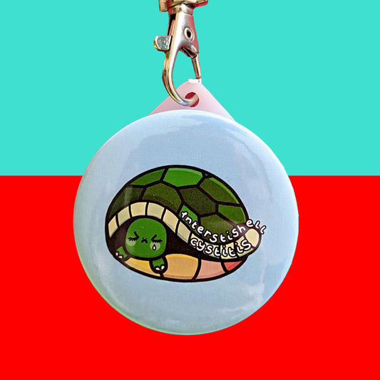 The Interstishell Cystitis Tortoise Keyring - IC Interstitial Cystitis on a red and blue background. The silver lobster clip circular keychain is a pale blue with a sad tortoise crying with white text reading 'interstishell cystitis'. The hand drawn design is raising awareness for Bladder pain syndrome interstitial cystitis.