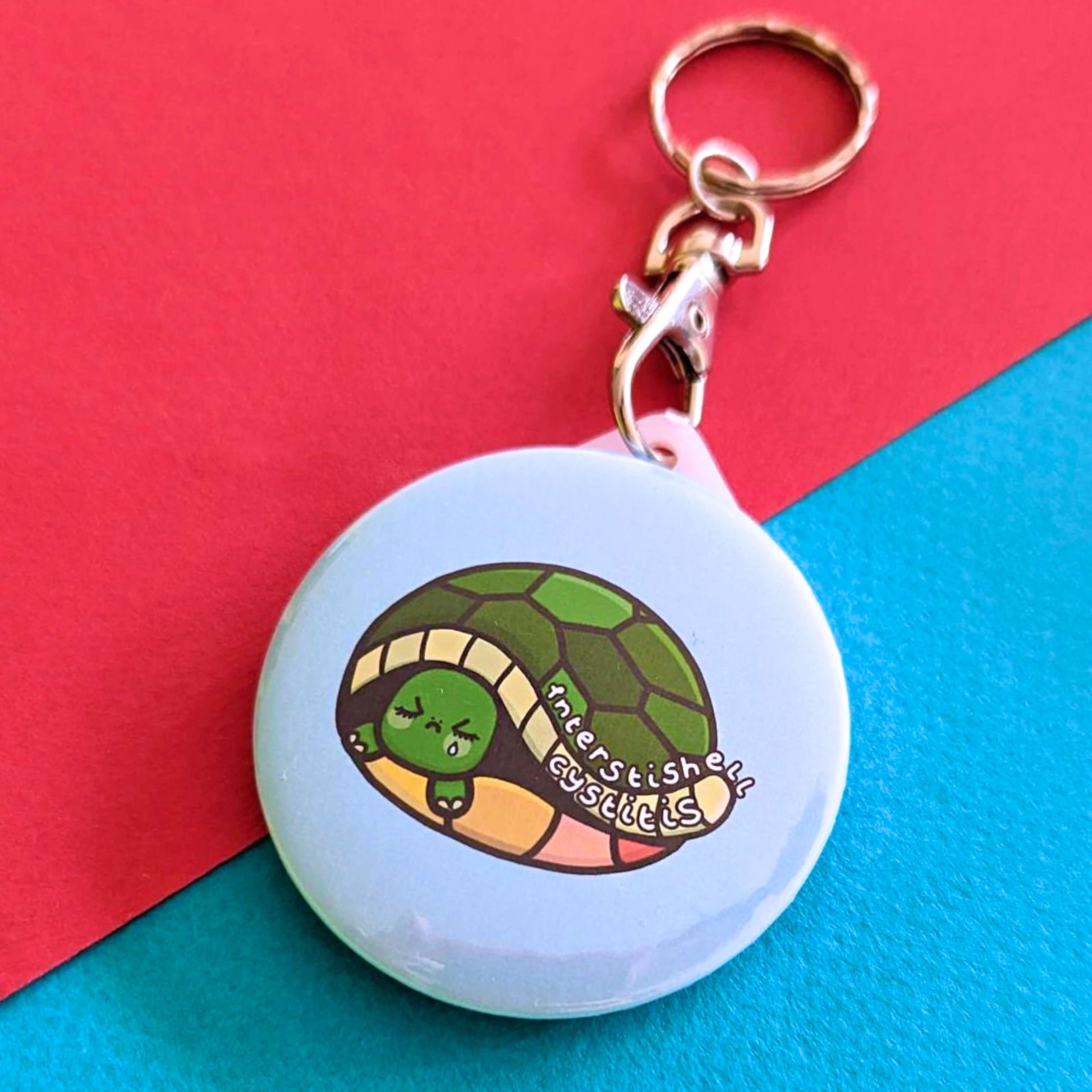 The Interstishell Cystitis Tortoise Keyring - IC Interstitial Cystitis on a red and blue background. The silver lobster clip circular keychain is a pale blue with a sad tortoise crying with white text reading 'interstishell cystitis'. The hand drawn design is raising awareness for Bladder pain syndrome interstitial cystitis.