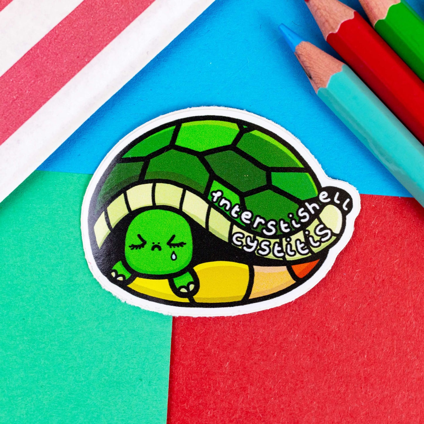 IC Interstishell cystitis sticker - IC Interstitial Cystitis. The sticker is of a tortoise hiding in its shell looking sad with text saying interstishell cystitis on its shell.  It is on a red, blue and green background with coloured pencils to one side and a stripy paper bag.