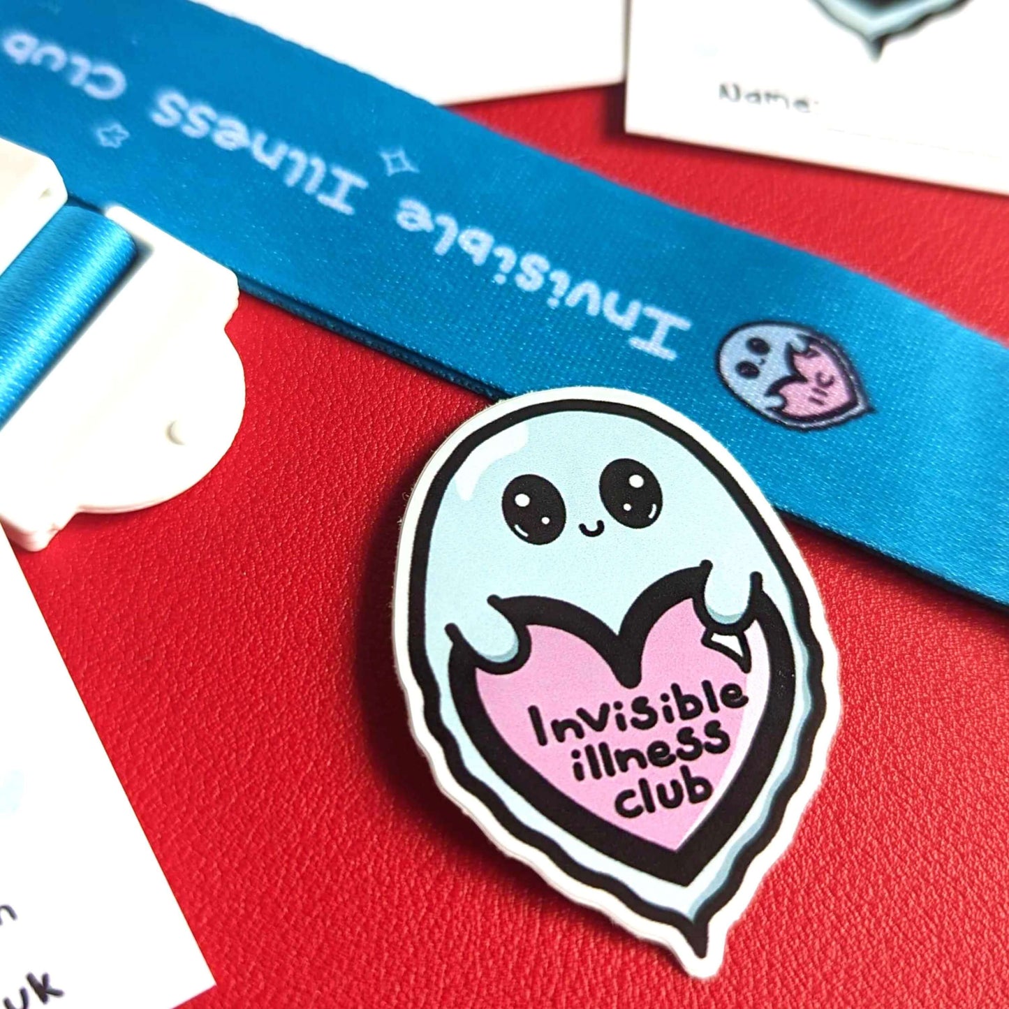 Invisible Illness Club vinyl sticker shown on red background. The sticker is in the shape of a cute mint green ghost with big eyes and little smile holding a pink heart with it's little hands that has 'invisible illness club' written in black writing in the middle of the heart.