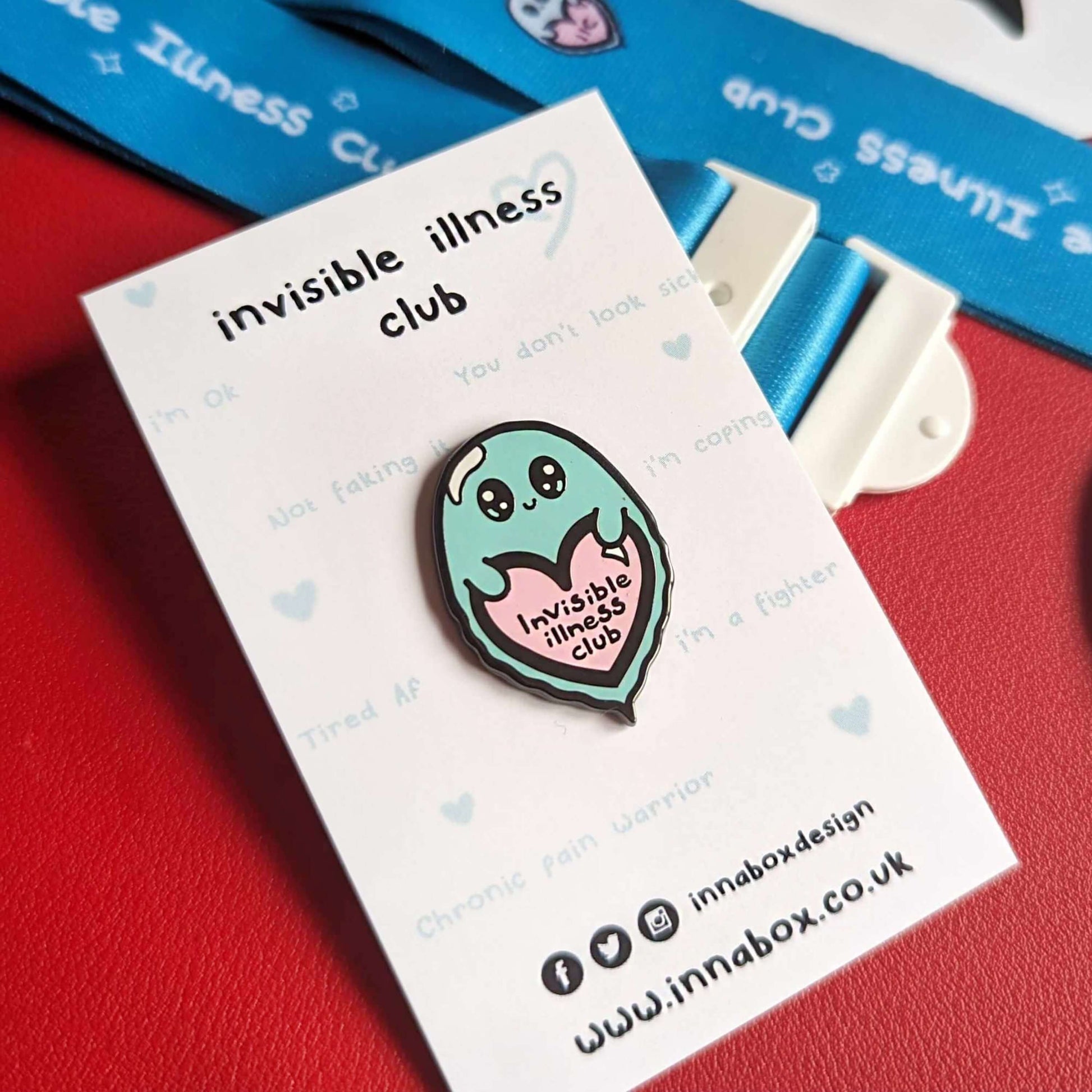 Invisible Illness Club enamel pin attached to it's backing card which has 'invisible illness club' written at the top and Innabox social media details at the bottom, shown on red background. The enamel pin is in the shape of a cute mint green ghost with big eyes and little smile holding a pink heart with it's little hands that has 'invisible illness club' written in black writing in the middle of the heart.