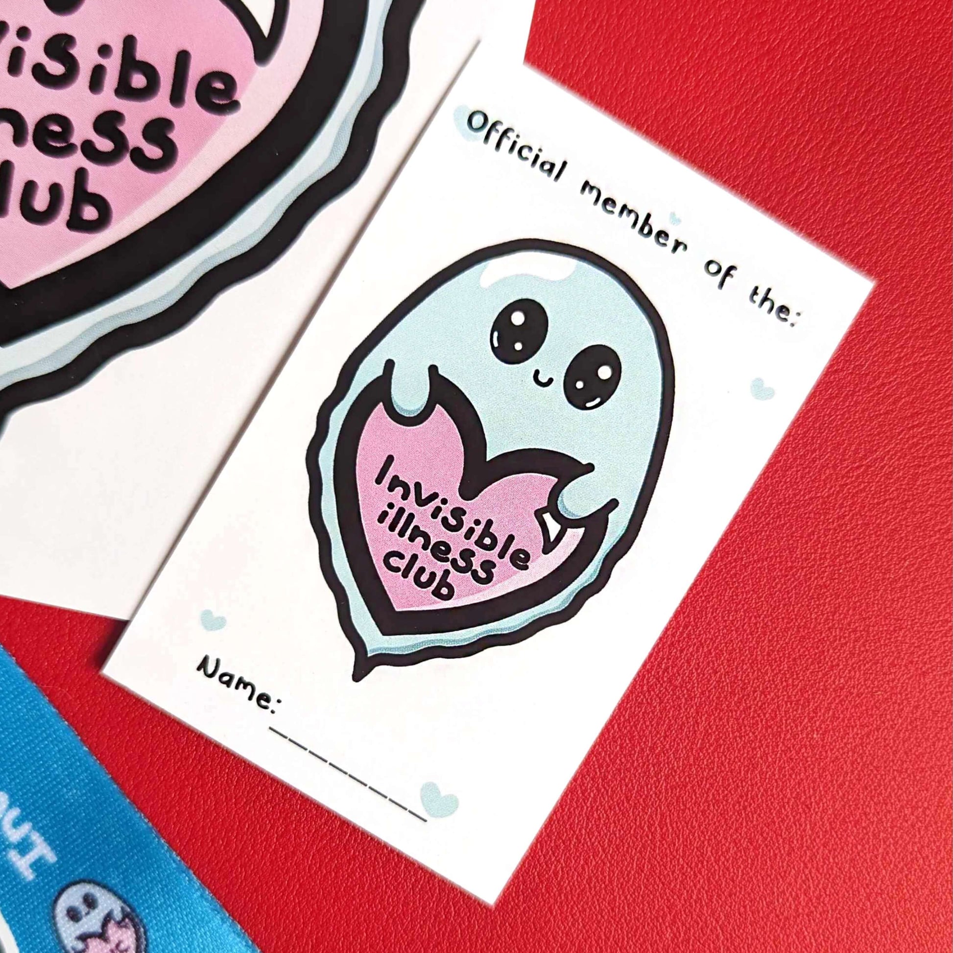 Close up of Invisible Illness Membership Card, a white card with little mint coloured hearts with 'official member of the:' written at the top in black writing and an illustration of a cute mint green ghost with big eyes and little smile holding a pink heart with it's little hands that has 'invisible illness club' written in black writing in the middle of the heart underneath. Underneath the ghost there is a space to write your name on the membership card. 