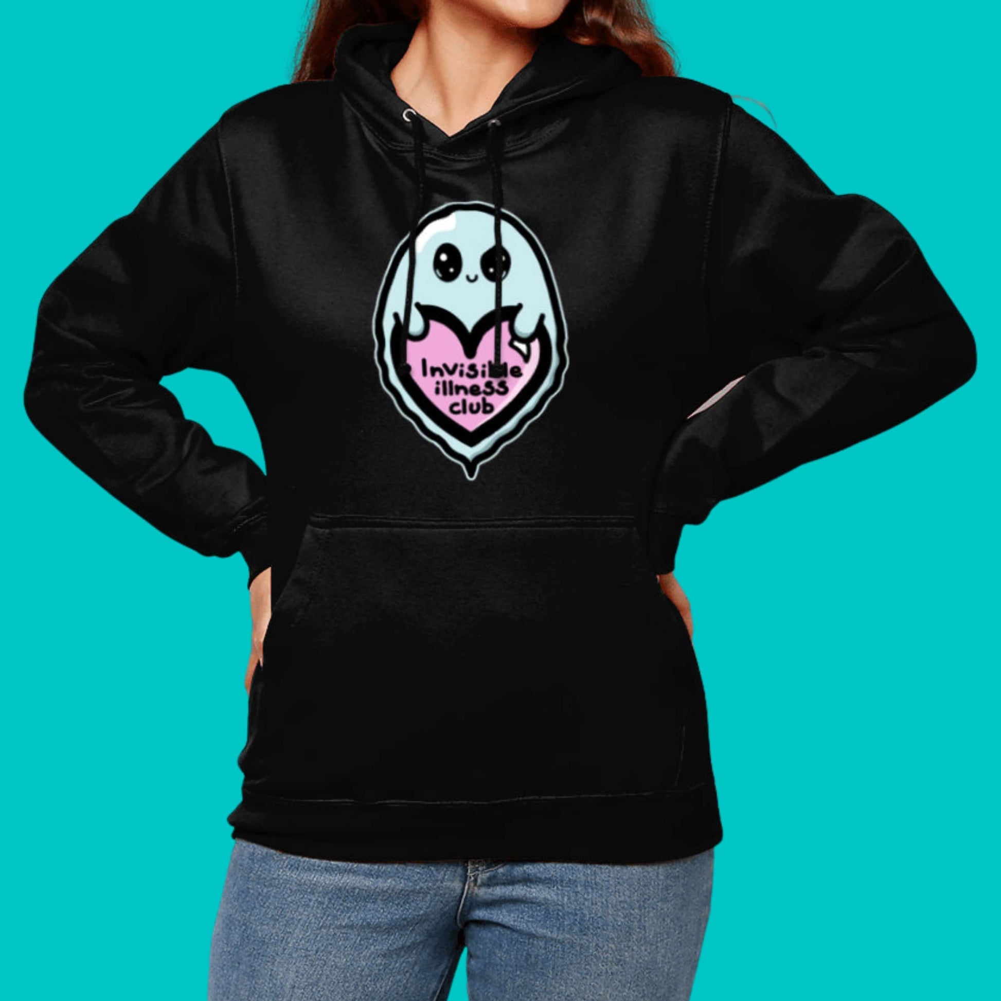 The Invisible Illness Club Black Hoodie modelled on a femme person with brown hair pairing the hoodie with blue denim jeans on a blue background. The black hoodie has a pastel blue smiling ghost with big sparkly eyes holding up a pastel pink heart with black text reading 'invisible illness club'. The hoodie has a large front pocket and black drawstring hood. The hand drawn design is raising awareness for hidden disabilites.
