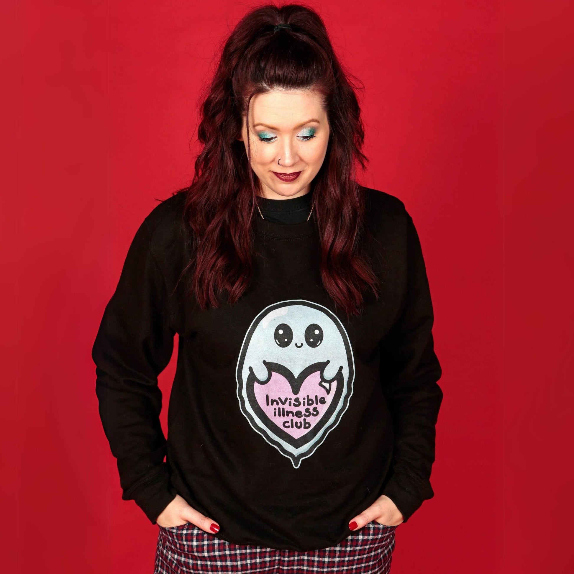 The Invisible Illness Club Sweatshirt Jumper being modelled by Nikky (owner of innabox) who has brown hair, blue eye makeup and blue and red nails. She is facing forward smiling looking down with both hands tucked in her red check trouser pockets. The black sweater features a smiling pastel blue ghost with big sparkly eyes holding up a pastel pink heart with black text reading 'invisible illness club'. The hand drawn design is raising awareness for hidden disabilities.