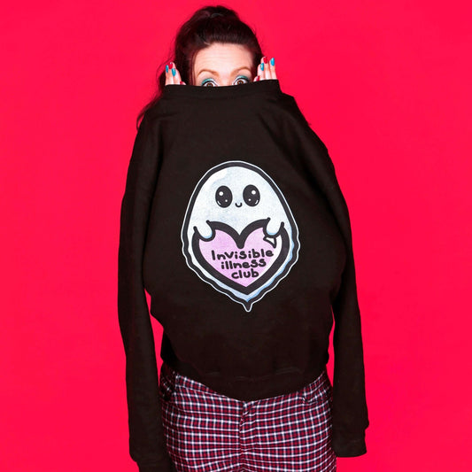 The Invisible Illness Club Sweatshirt Jumper being modelled by Nikky (owner of innabox) who has brown hair, blue eye makeup and blue and red nails. She is facing forward poking out the black sweatshirt jumper top with a shocked wide eyed expression, paired with red check trousers. The black sweater features a smiling pastel blue ghost with big sparkly eyes holding up a pastel pink heart with black text reading 'invisible illness club'. The hand drawn design is raising awareness for hidden disabilities.