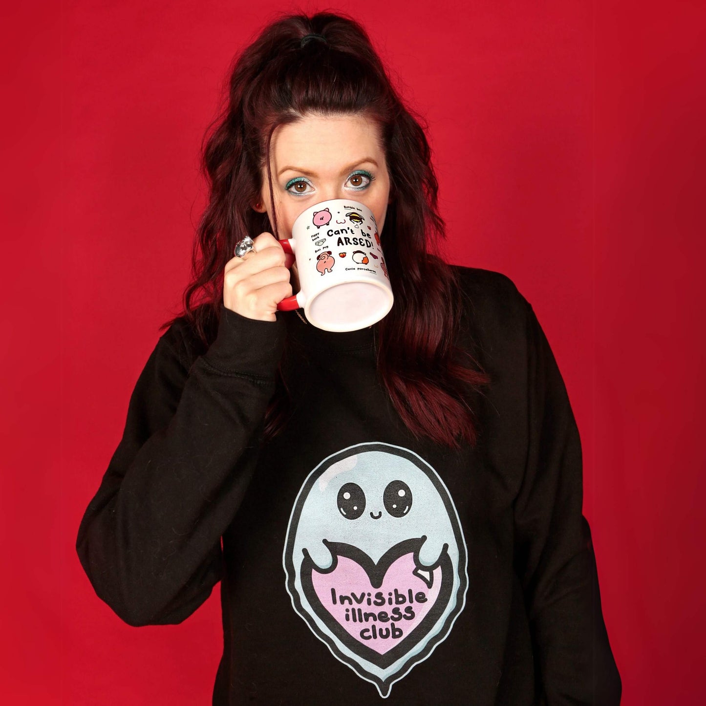 The Invisible Illness Club Sweatshirt Jumper being modelled by Nikky (owner of innabox) who has brown hair, blue eye makeup and blue and red nails. She is facing forward lifting the innabox can't be arsed mug to her face. The black sweater features a smiling pastel blue ghost with big sparkly eyes holding up a pastel pink heart with black text reading 'invisible illness club'. The hand drawn design is raising awareness for hidden disabilities.