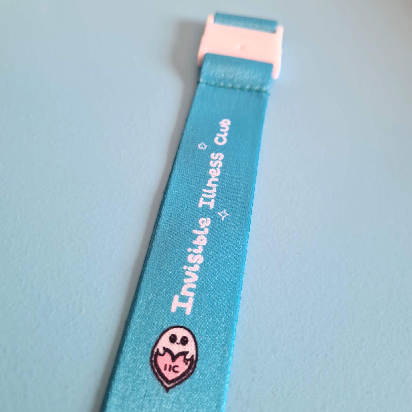 The Invisible Illness Club Lanyard on a pastel blue background. The pastel blue lanyard has repeating white text reading 'invisible illness club' and innabox pastel ghost logo. It has a silver lobster clip and white safety break. The hand drawn design is raising awareness for hidden disabilities.