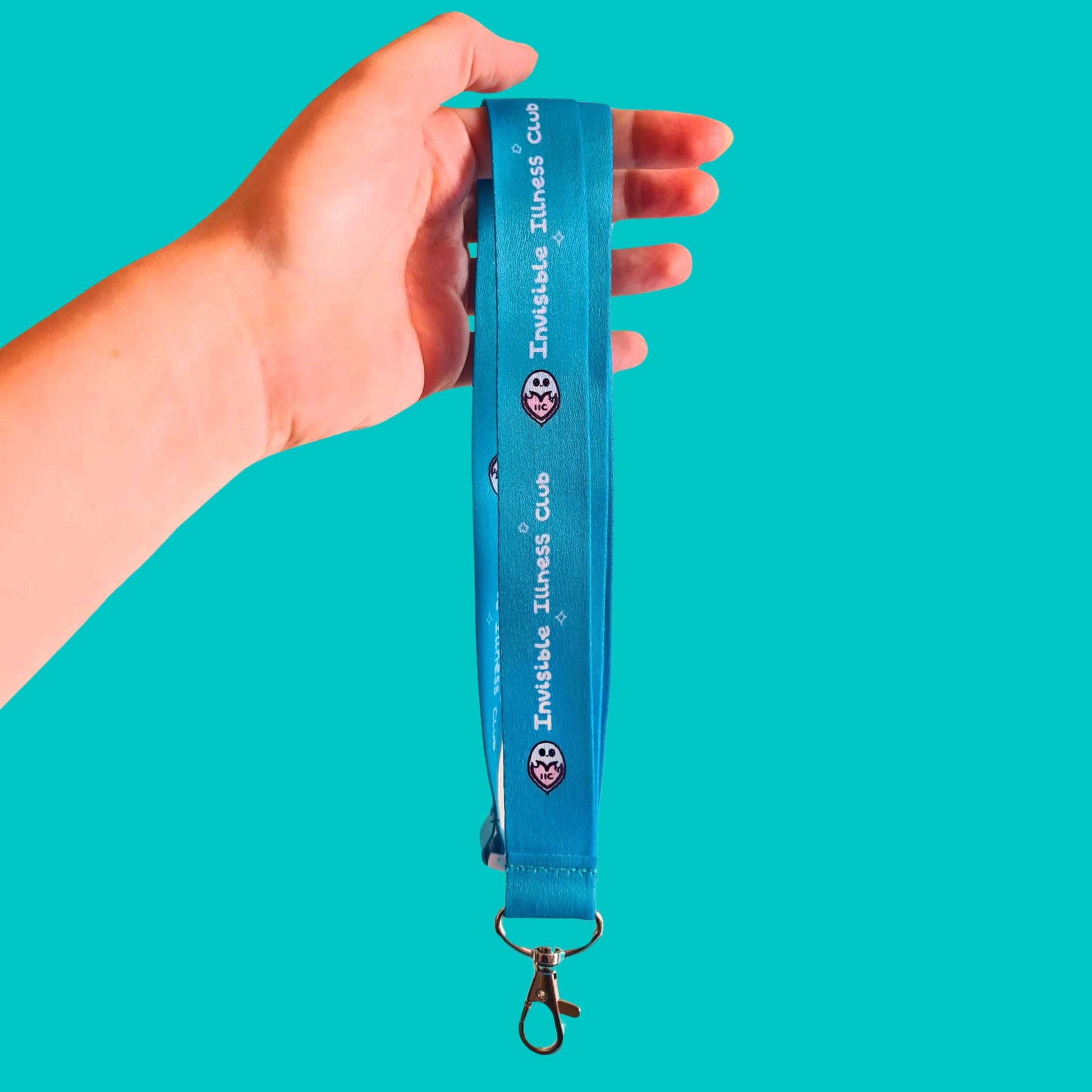 The Invisible Illness Club Lanyard held over a blue background. The pastel blue lanyard has repeating white text reading 'invisible illness club' and innabox pastel ghost logo. It has a silver lobster clip and white safety break. The hand drawn design is raising awareness for hidden disabilities.