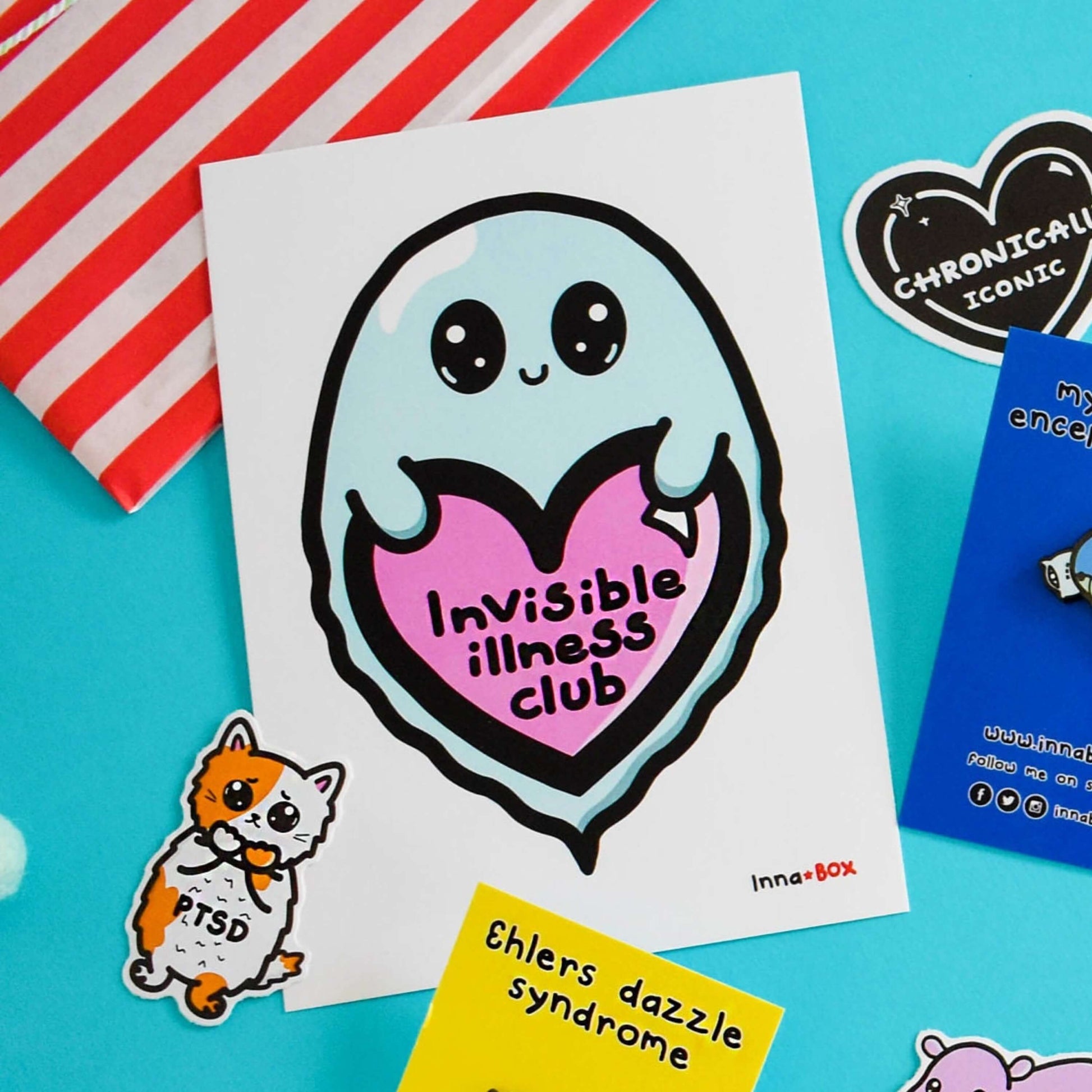 The Invisible Illness Club Postcard on a blue background with other innabox products. The white postcard print features a pastel blue smiling ghost with big sparkly eyes holding up a pink heart with black text reading 'invisible illness club' with the innabox logo in the bottom right corner. The hand drawn design is raising awareness for hidden disabilities and chronic illness.