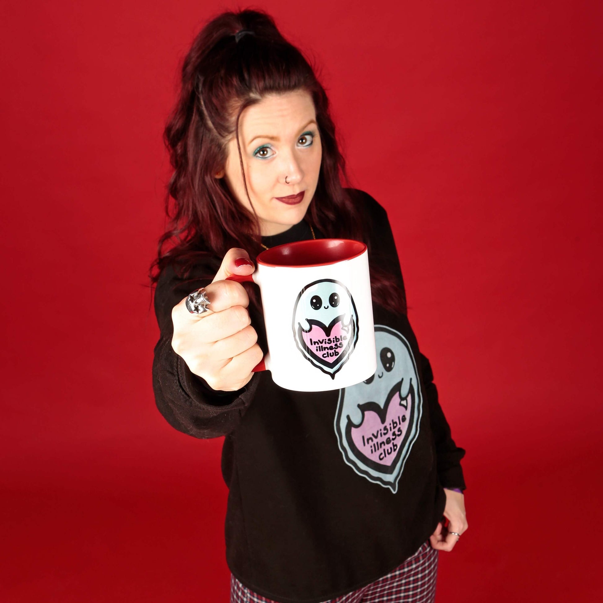 The Invisible Illness Club Mug being held up by Nikky, who owns innabox, wearing the invisible illness club black sweater and red check trousers. The white mug with red handle and inside has an illustration of a cute smiling ghost holding a pink heart with text saying invisible illness club. Hand drawn design made to raise awareness for hidden and chronic illnesses.