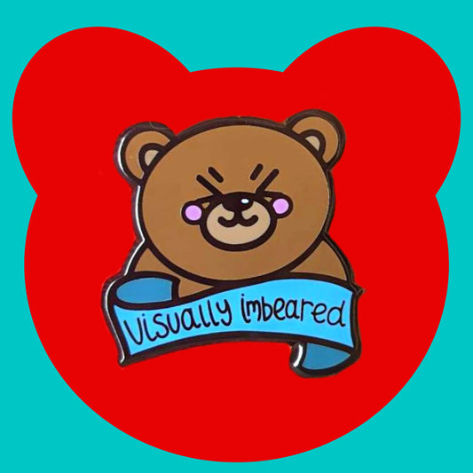 The Visually Imbeared Bear Enamel Pin - Visually Impaired on a red and blue background. The brown teddy bear shaped pin has its eyes scrunched shut smiling with pink cheeks, underneath is a blue banner with black text reading 'visually imbeared'. The hand drawn design is raising awareness for visual impairment and blindness.