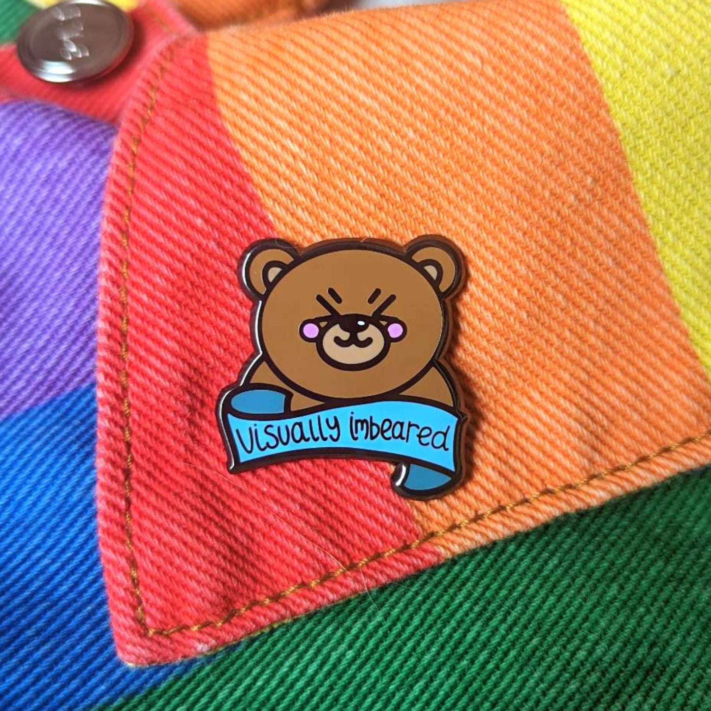 The Visually Imbeared Bear Enamel Pin - Visually Impaired on a rainbow denim jacket collar. The brown teddy bear shaped pin has its eyes scrunched shut smiling with pink cheeks, underneath is a blue banner with black text reading 'visually imbeared'. The hand drawn design is raising awareness for visual impairment and blindness.