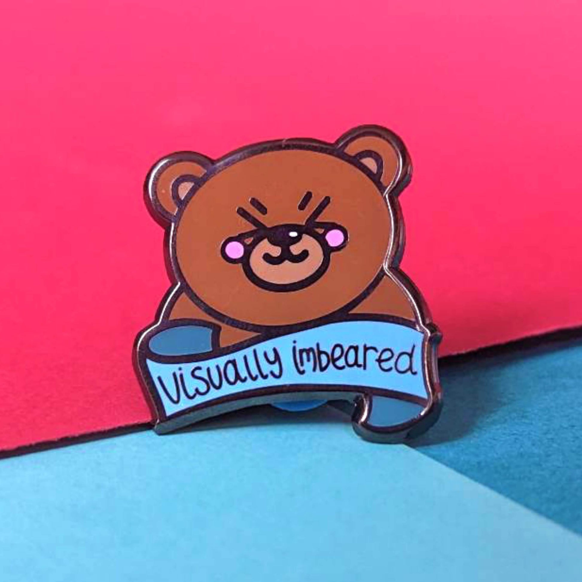 The Visually Imbeared Bear Enamel Pin - Visually Impaired on a red and blue background. The brown teddy bear shaped pin has its eyes scrunched shut smiling with pink cheeks, underneath is a blue banner with black text reading 'visually imbeared'. The hand drawn design is raising awareness for visual impairment and blindness.