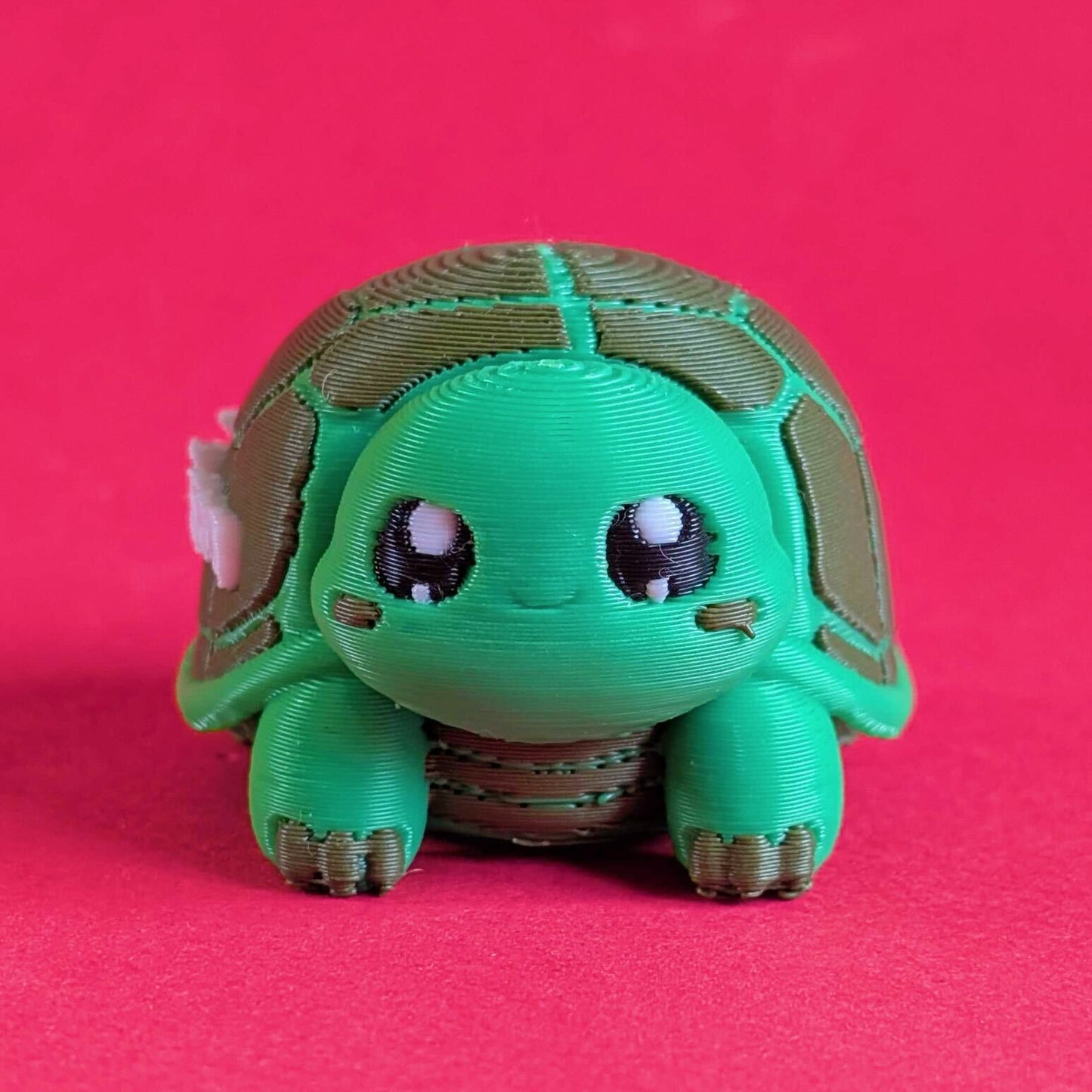 Front facing view of a green 3D printed tortoise desk buddy fidget toy with gentle expression and chunky feet. Tortism Tortoise Desk Buddy by Innabox, a calming sensory object designed for autistic people and hidden disabilities, photographed on a bright pink background.
