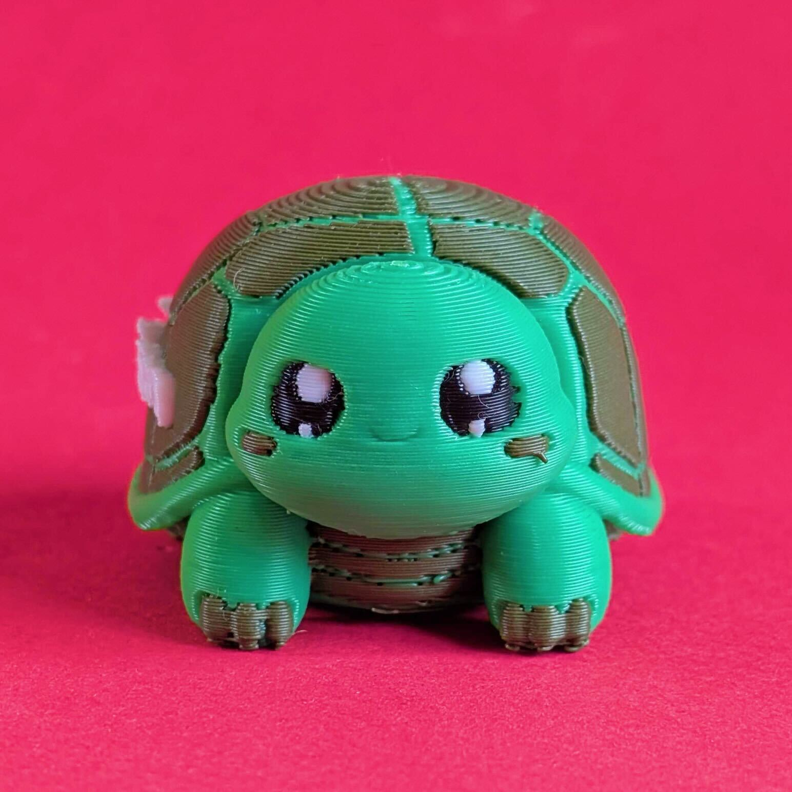 Front facing view of a green 3D printed tortoise desk buddy fidget toy with gentle expression and chunky feet. Tortism Tortoise Desk Buddy by Innabox, a calming sensory object designed for autistic people and hidden disabilities, photographed on a bright pink background.
