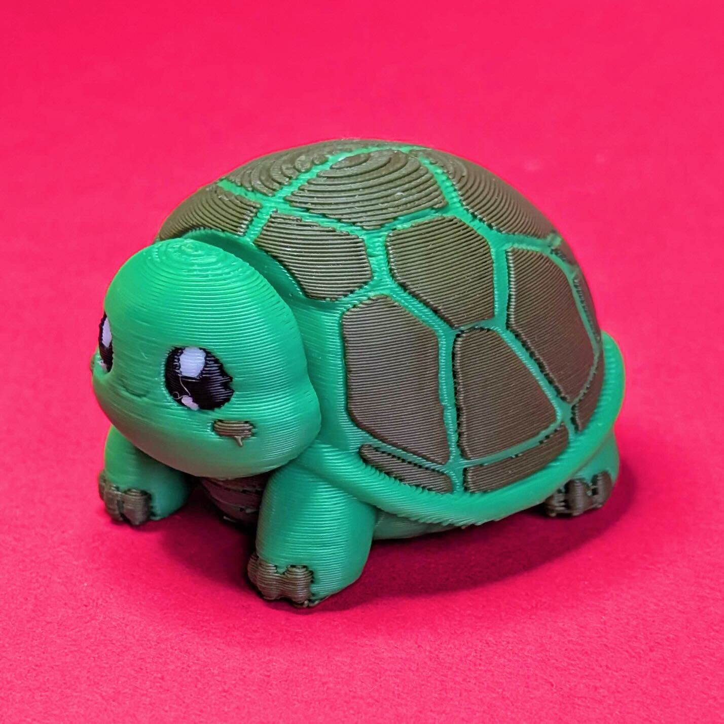 Side view of a green 3D printed tortoise desk buddy fidget toy with rounded legs and textured shell. Tortism Tortoise Desk Buddy by Innabox, a comforting sensory companion designed for autistic people and those with chronic illness, photographed on a pink background.