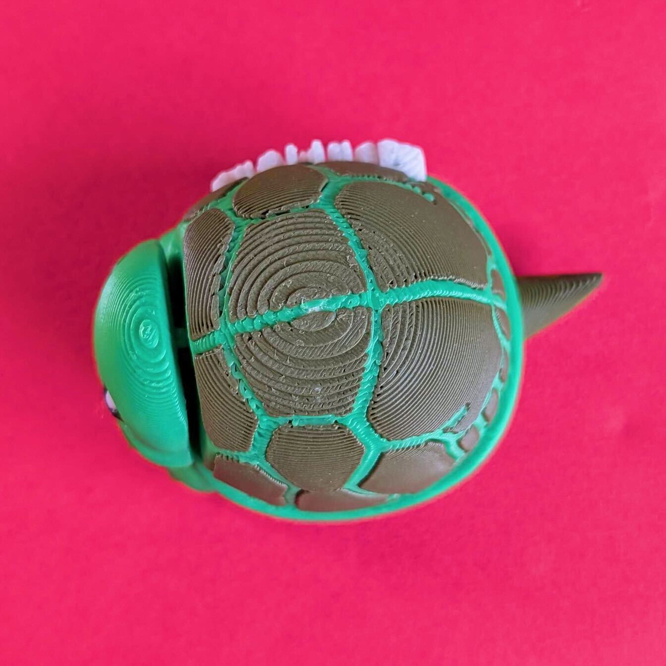Top down view of a green 3D printed tortoise fidget toy showing detailed shell texture and layered design. Tortism Tortoise Desk Buddy by Innabox, a small autism friendly sensory desk buddy made from biodegradable PLA, photographed on a bright pink background.