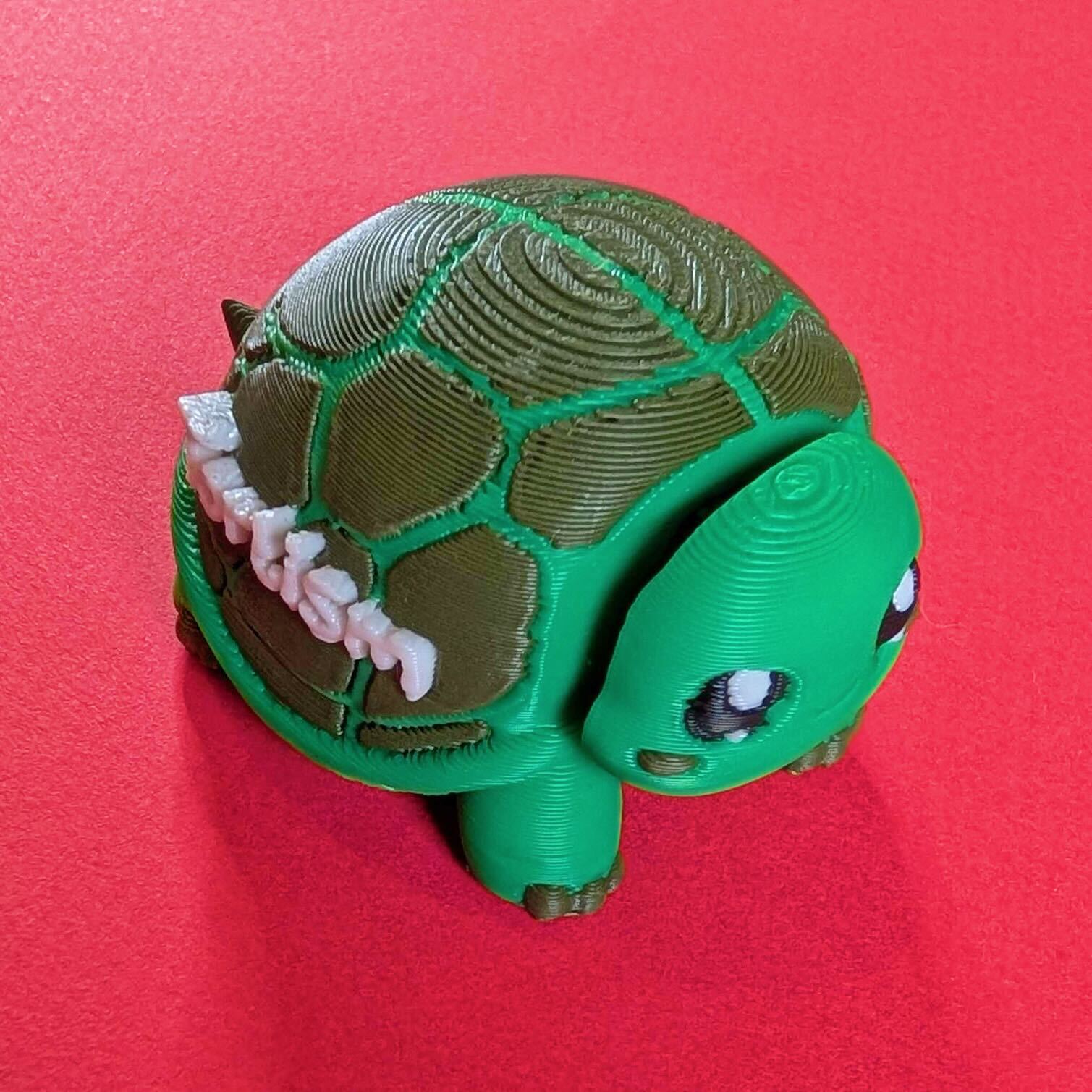 Green 3D printed tortoise desk buddy fidget toy with textured shell and cute black and white eyes, photographed on a bright pink background. Tortism Tortoise Desk Buddy by Innabox, a small sensory autism friendly fidget designed to sit in the palm of your hand or on a desk, made from biodegradable polylactic acid.