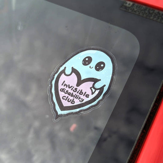 The Invisible Disability Club Ghost Window Cling on a red car window. The pastel blue smiling Ghost window sticker has big sparkly eyes holding a pastel pink heart with black text reading 'invisible disability club'. The hand drawn design is raising awareness for hidden disabilities and invisible illnesses.