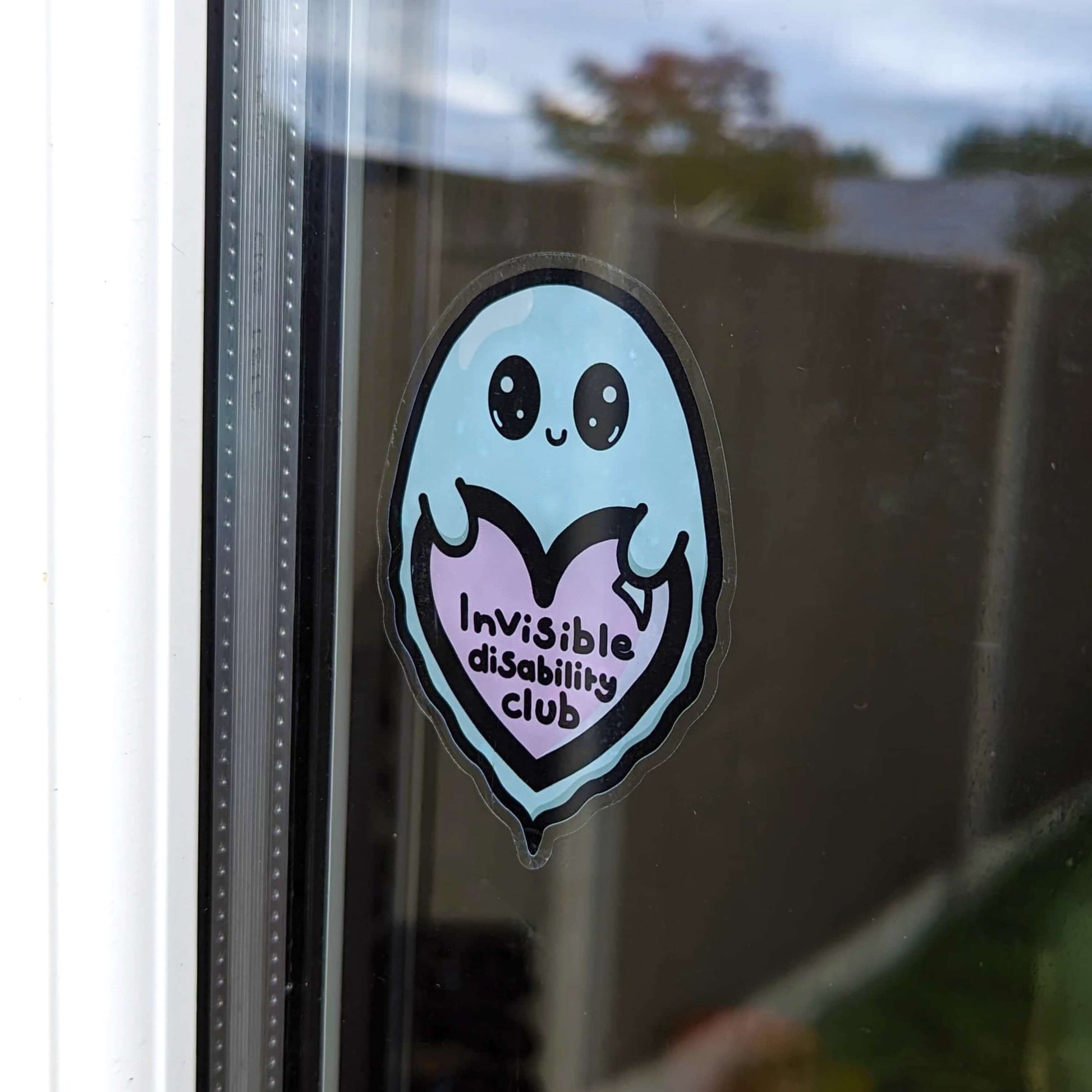  The Invisible Disability Club Ghost Window Cling on a house back door window. The pastel blue smiling Ghost window sticker has big sparkly eyes holding a pastel pink heart with black text reading 'invisible disability club'. The hand drawn design is raising awareness for hidden disabilities and invisible illnesses.