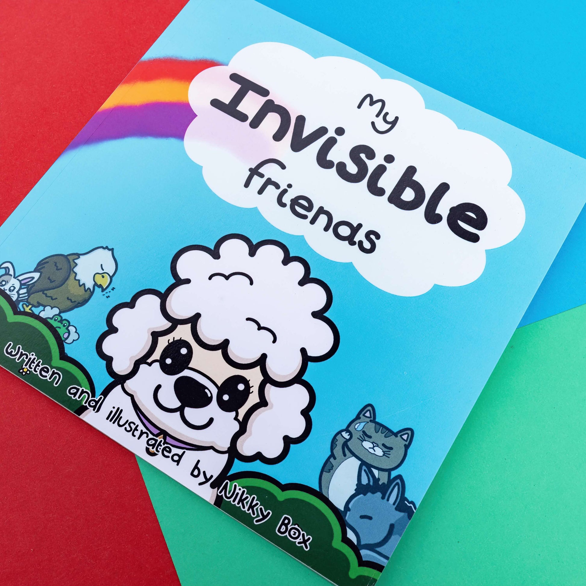 The My Invisible Friends Children's Book on a red, blue and green background. The blue book features a white smiling poodle with disabled innabox characters in the background with a rainbow and white cloud above with black text reading 'my invisible friends' and bottom black text reading 'written and illustrated by Nikky box'. Raising awareness for invisible illnesses.