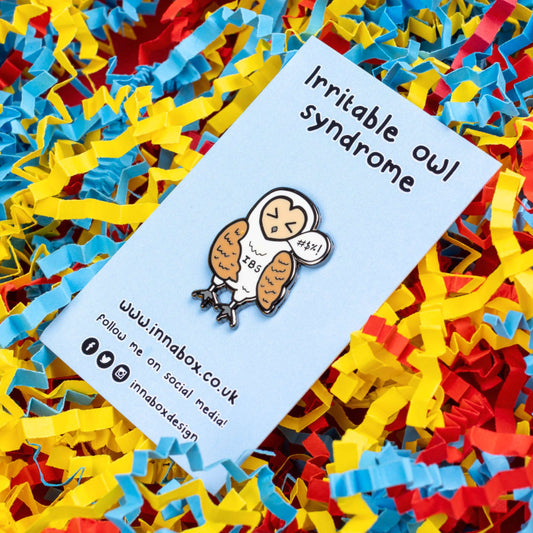 Irritable Owl Syndrome Enamel Pin - Irritable bowel syndrome (IBS) on pastel blue backing card on a red, yellow and blue card confetti background. The enamel pin is of a barn owl with a sweary text speech bubble and has its eyes shut with the letters IBS written on its belly. Hand drawn design to raise awareness for Irritable bowel syndrome (IBS).
