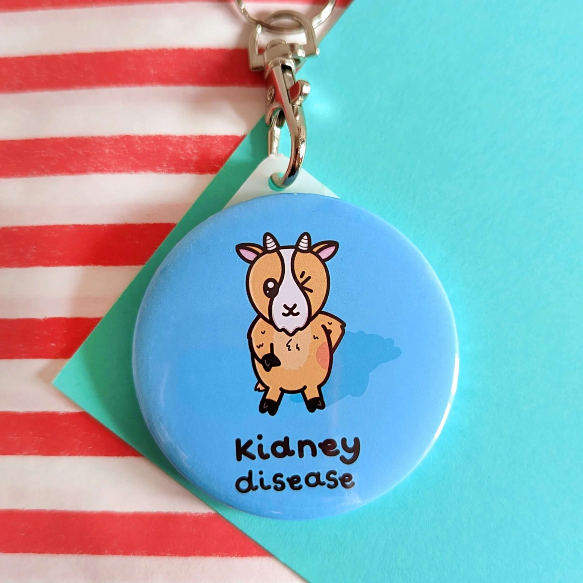 The Kidney Disease Goat Keyring - Kidney Disease on a red and blue background. The pastel blue plastic circular keychain has a silver lobster clip and an orange goat with little horns stood up winking with a red right hand side, underneath in black text reads 'kidney disease'. The hand drawn design is raising awareness for kidney disease and kidney failure.
