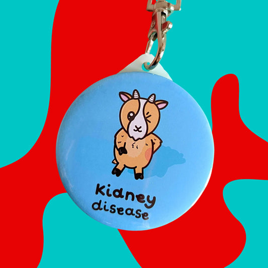 The Kidney Disease Goat Keyring - Kidney Disease on a red and blue background. The pastel blue plastic circular keychain has a silver lobster clip and an orange goat with little horns stood up winking with a red right hand side, underneath in black text reads 'kidney disease'. The hand drawn design is raising awareness for kidney disease and kidney failure.