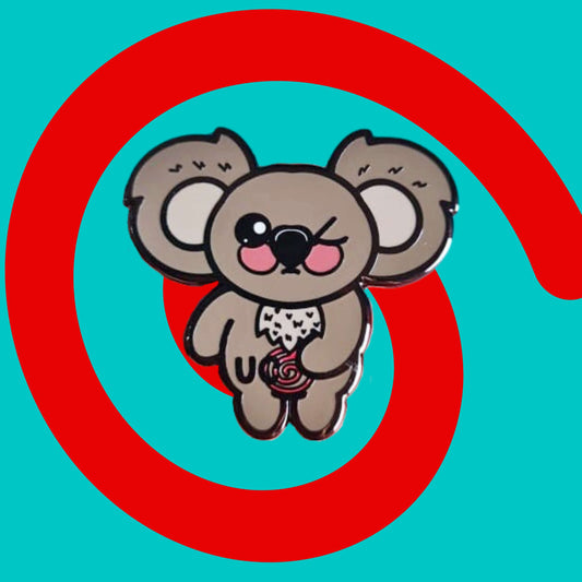 The Ulcerative Koalitis Koala Enamel Pin - Ulcerative Colitis UC on a red and blue background. The grey koala shaped enamel pin has one eye shut clutching its red swirly tummy with black text over it reading 'UC'. The hand drawn design is raising awareness for Ulcerative Colitis UC.