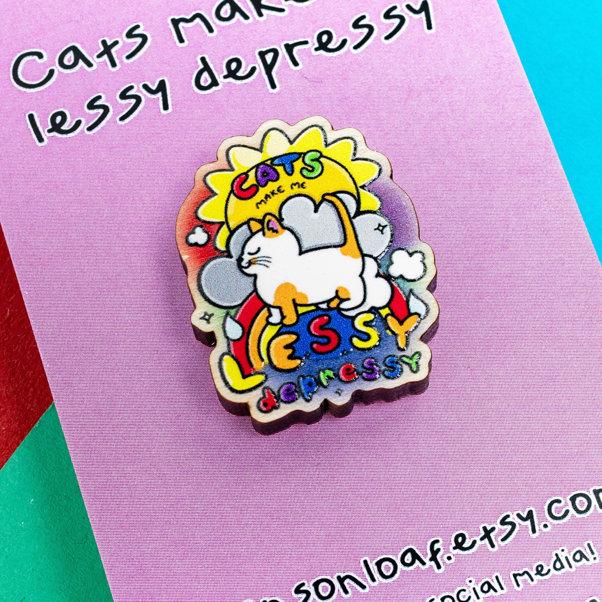 Close up of the Cats Make Me Lessy Depressy Wooden Pin Badge on pink backing card laying on a red, blue and green card background. The wooden pin features a smiling orange and white cat with a sunshine, raincloud and rainbow, in rainbow text across this reads 'cats make me lessy depressy'. Hand drawn design to raise awareness for mental health and depression.