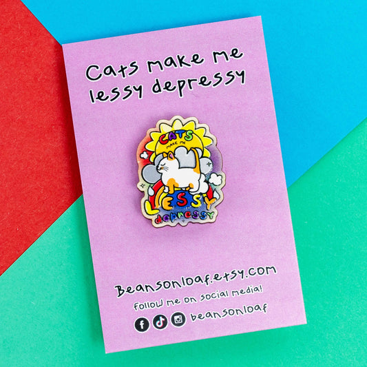 The Cats Make Me Lessy Depressy Wooden Pin Badge on pink backing card laying on a red, blue and green card background. The wooden pin features a smiling orange and white cat with a sunshine, raincloud and rainbow, in rainbow text across this reads 'cats make me lessy depressy'. Hand drawn design to raise awareness for mental health and depression.