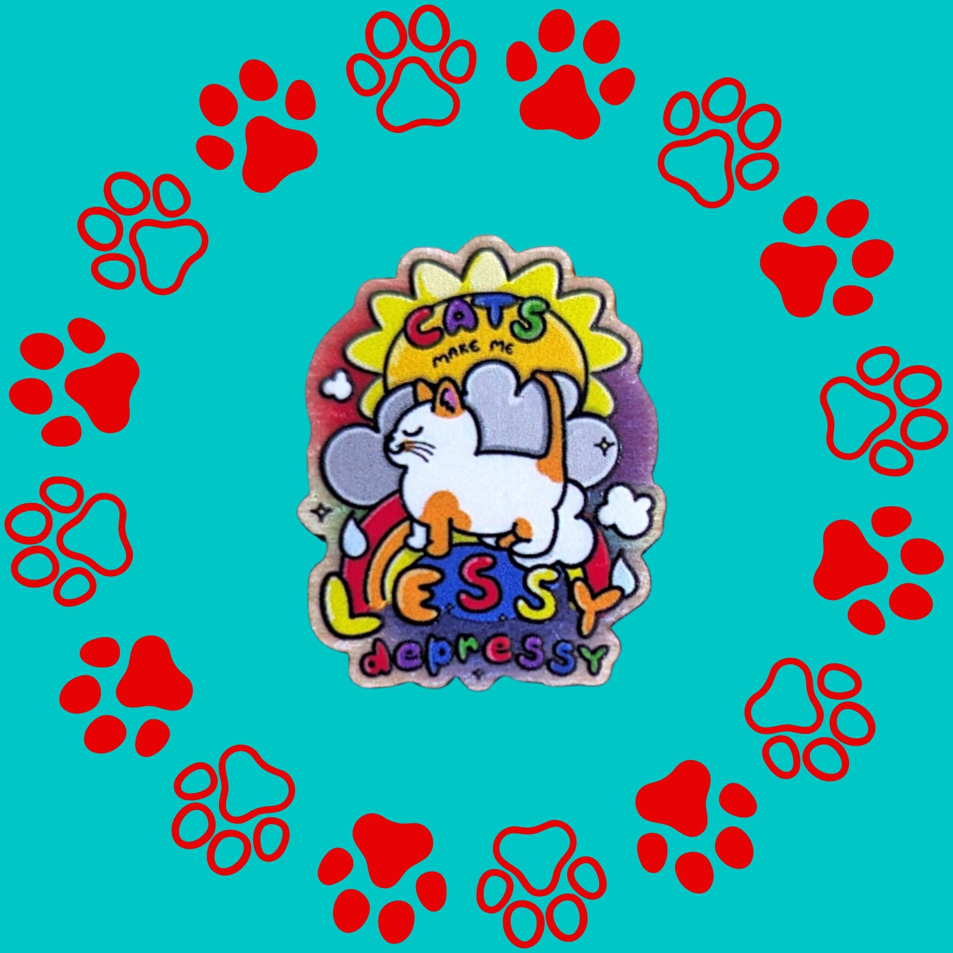 The Cats Make Me Lessy Depressy Wooden Pin Badge on a red and blue paw print background. The wooden pin features a smiling orange and white cat with a sunshine, raincloud and rainbow, in rainbow text across this reads 'cats make me lessy depressy'. Hand drawn design to raise awareness for mental health and depression.