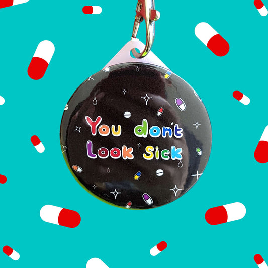 The You Don't Look Sick Keyring on a blue background with red and white pills all over. The silver lobster clip black plastic circular keyring has rainbow writing reading 'you don't look sick' with colourful pills and white sparkles all over.