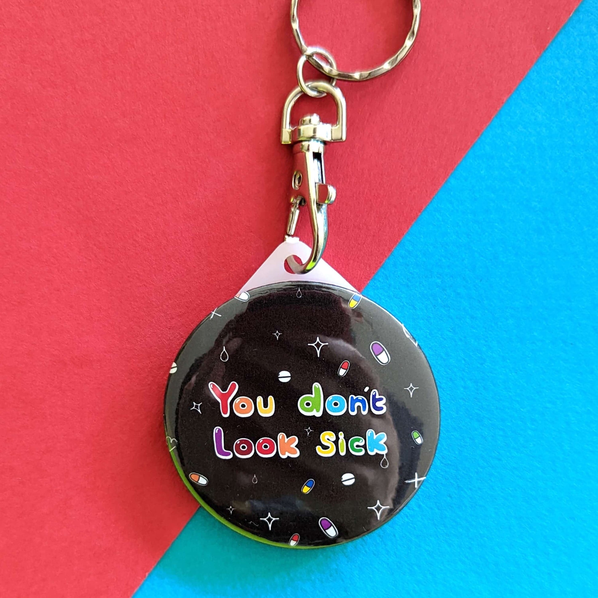 The You Don't Look Sick Keyring on a red and blue background. The silver lobster clip black plastic circular keyring has rainbow writing reading 'you don't look sick' with colourful pills and white sparkles all over.