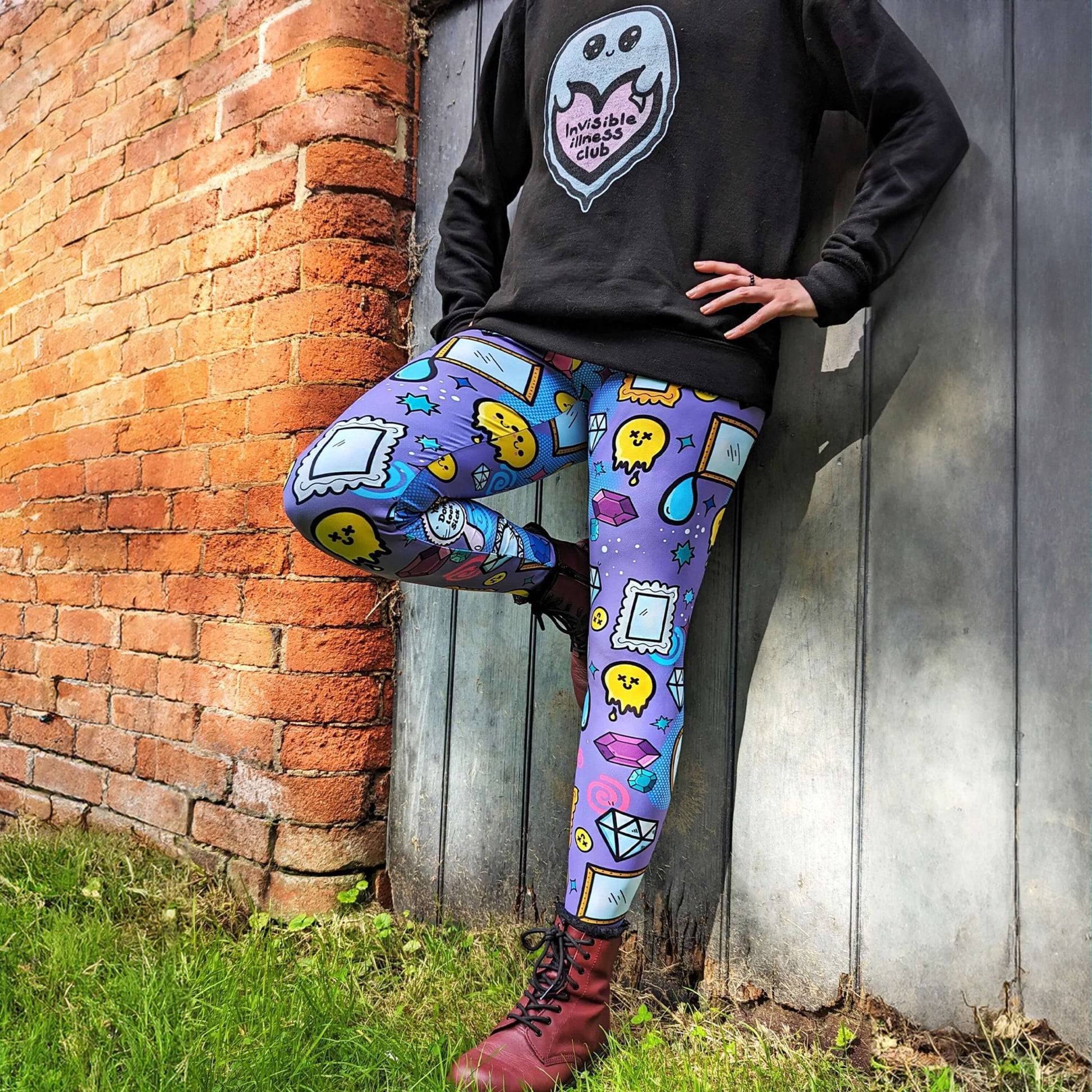 The You Don't Look Sick Leggings modelled by the owner of Innabox Nikky with red boots and the innabox invisible illness club sweater. She is leaning back on a wooden door next to a brick wall. The purple base leggings features melting yellow smiley faces, mirrors, gemstones, teardrops, sparkles, swirls and dots with a centre hand held mirror reading 'you don't look sick'. The hand drawn design is raising awareness for invisible illnesses.