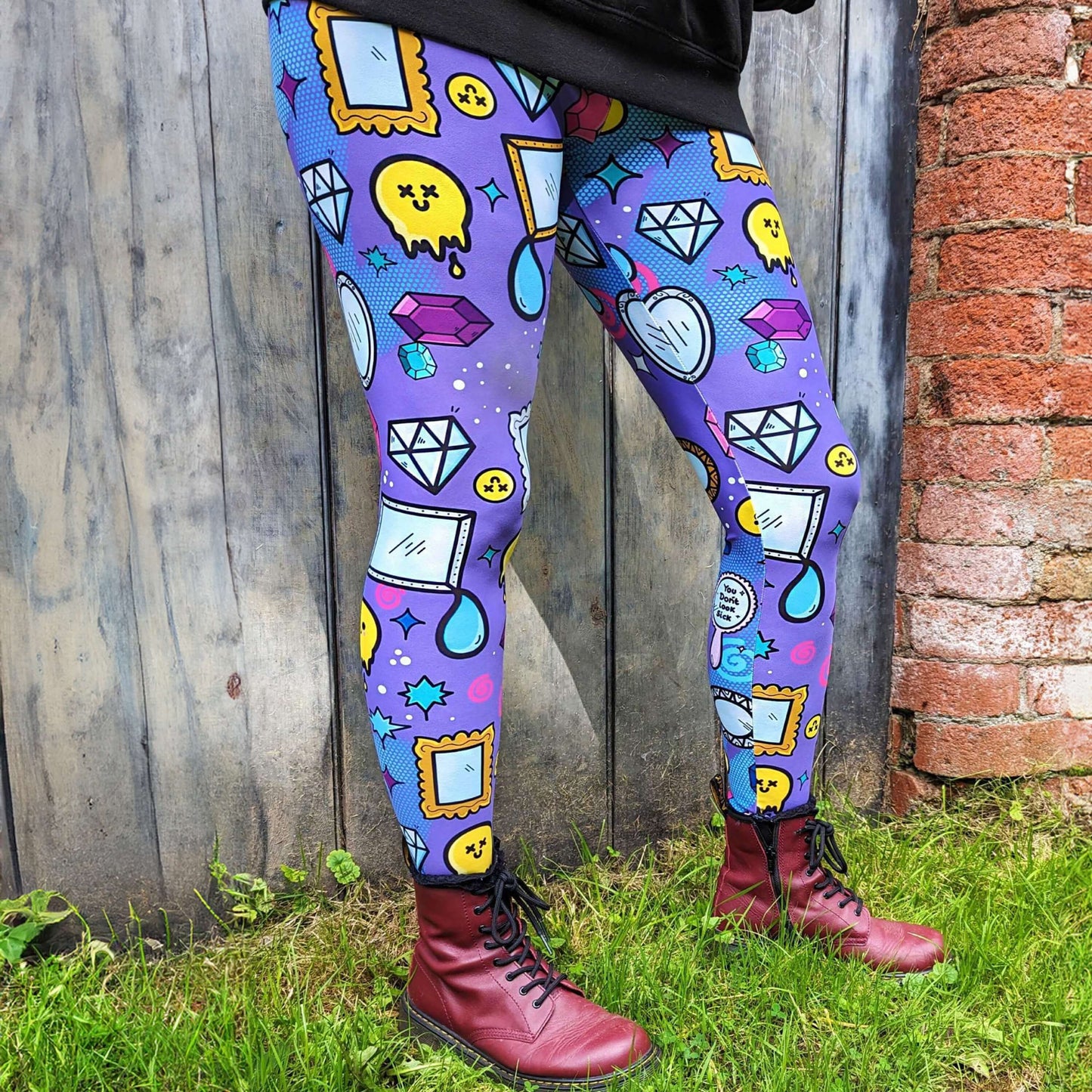 The You Don't Look Sick Leggings modelled by the owner of Innabox Nikky with red boots and the innabox invisible illness club sweater. She is stood in front of a wooden door next to a brick wall. The purple base leggings features melting yellow smiley faces, mirrors, gemstones, teardrops, sparkles, swirls and dots with a centre hand held mirror reading 'you don't look sick'. The hand drawn design is raising awareness for invisible illnesses.