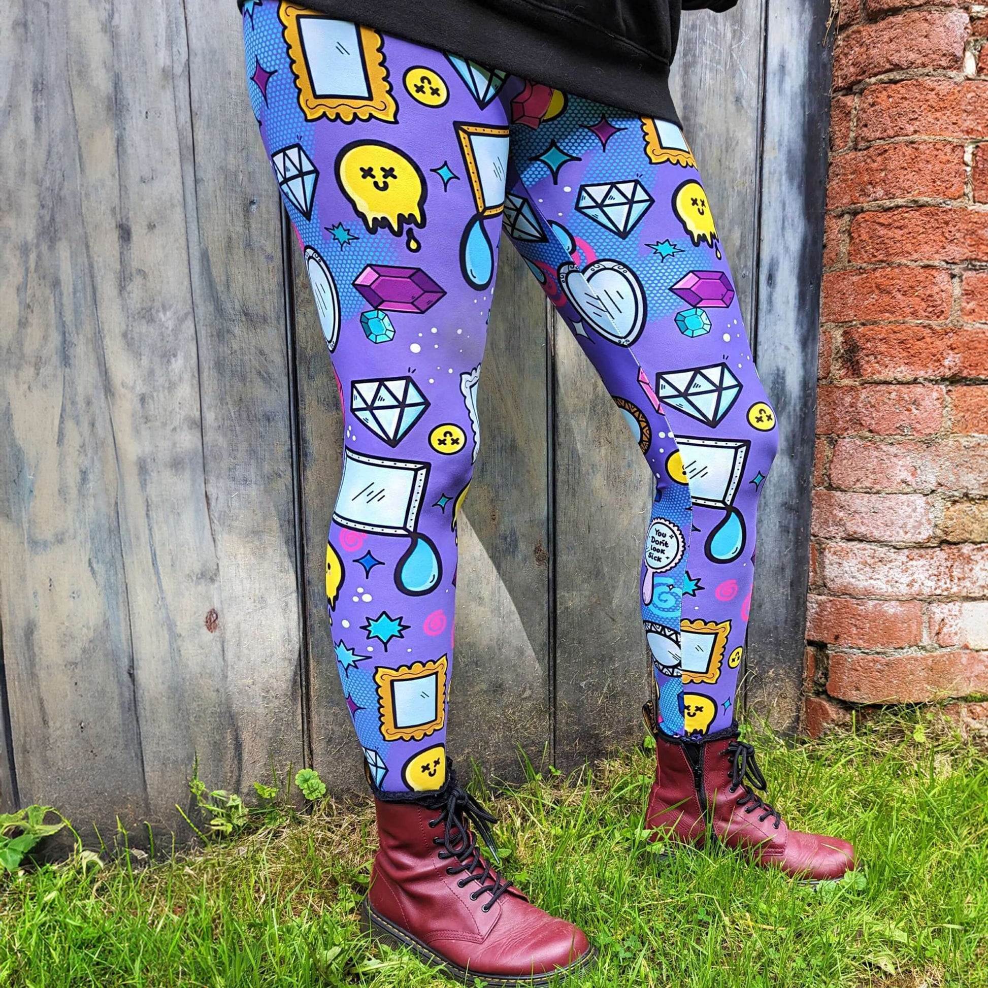 The You Don't Look Sick Leggings modelled by the owner of Innabox Nikky with red boots and the innabox invisible illness club sweater. She is stood in front of a wooden door next to a brick wall. The purple base leggings features melting yellow smiley faces, mirrors, gemstones, teardrops, sparkles, swirls and dots with a centre hand held mirror reading 'you don't look sick'. The hand drawn design is raising awareness for invisible illnesses.