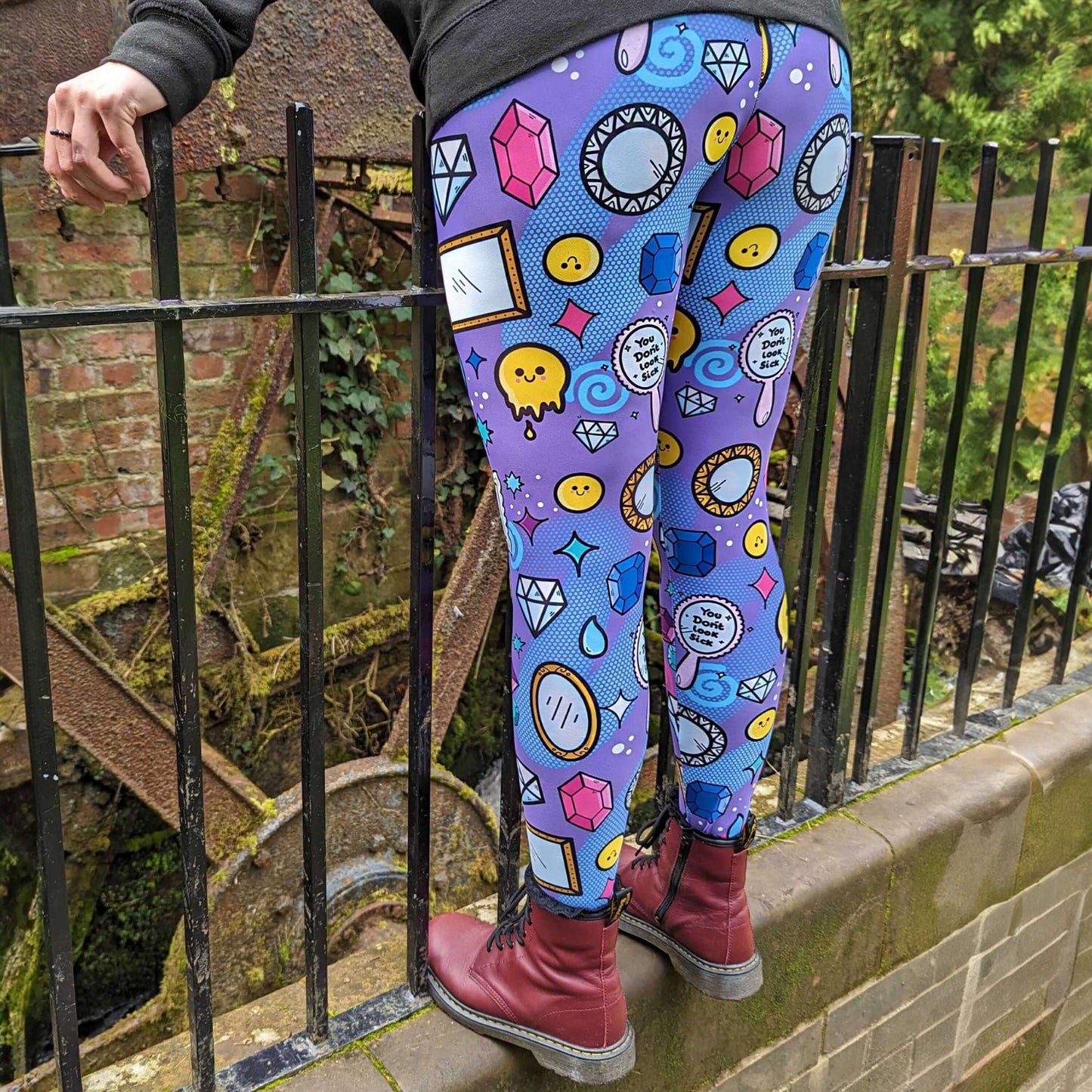 The You Don't Look Sick Leggings modelled by the owner of Innabox Nikky with red boots and the innabox invisible illness club sweater. She is stood leaning forward on a metal railing to highlight the leggings. The purple base leggings features melting yellow smiley faces, mirrors, gemstones, teardrops, sparkles, swirls and dots with a centre hand held mirror reading 'you don't look sick'. The hand drawn design is raising awareness for invisible illnesses.