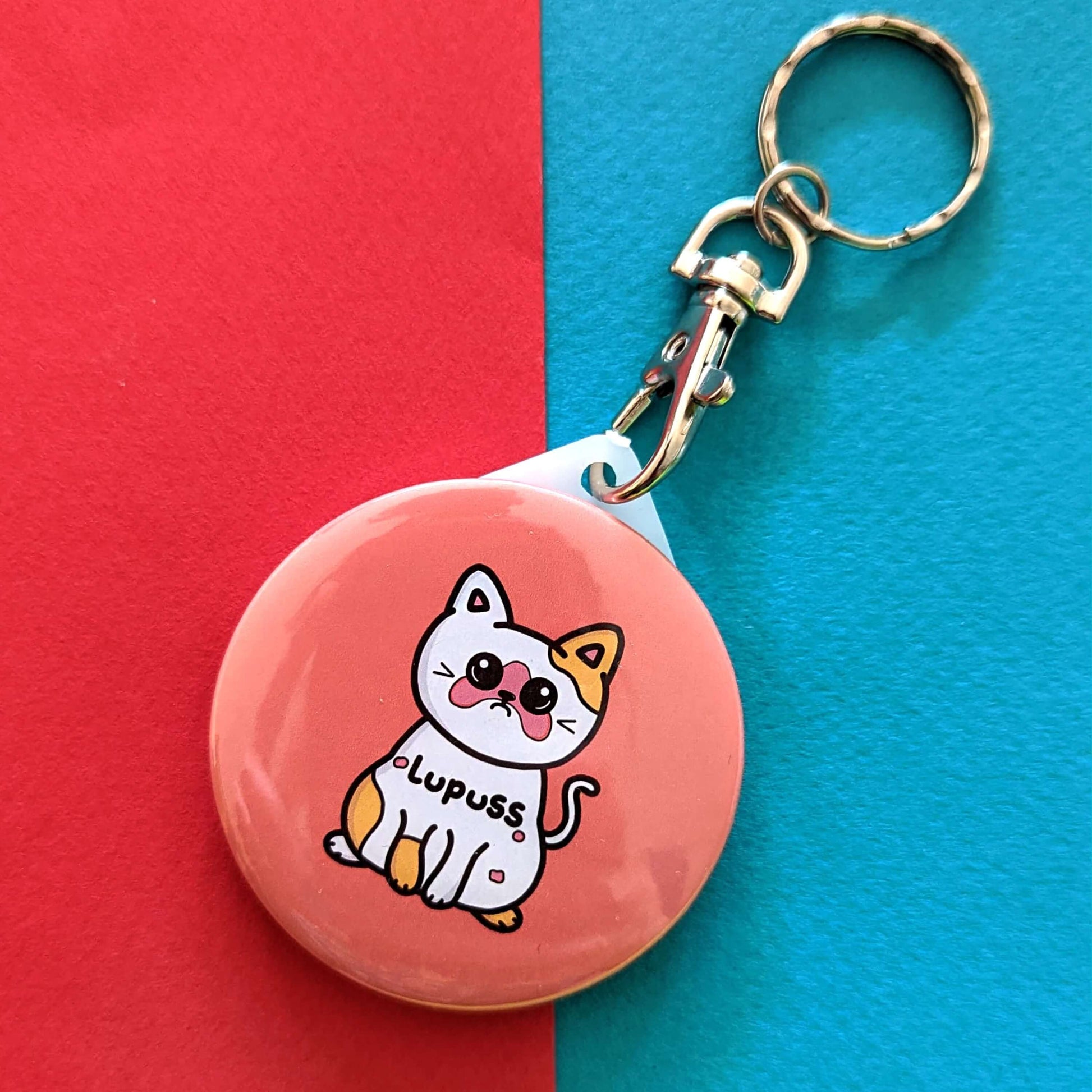 The Lupuss Cat Keyring - Lupus on a red and blue background. The peach pink circular keychain with silver lobster clip features a sad calico cat with red patches with black text across its body reading 'lupuss'. The hand drawn design is raising awareness for lupus.