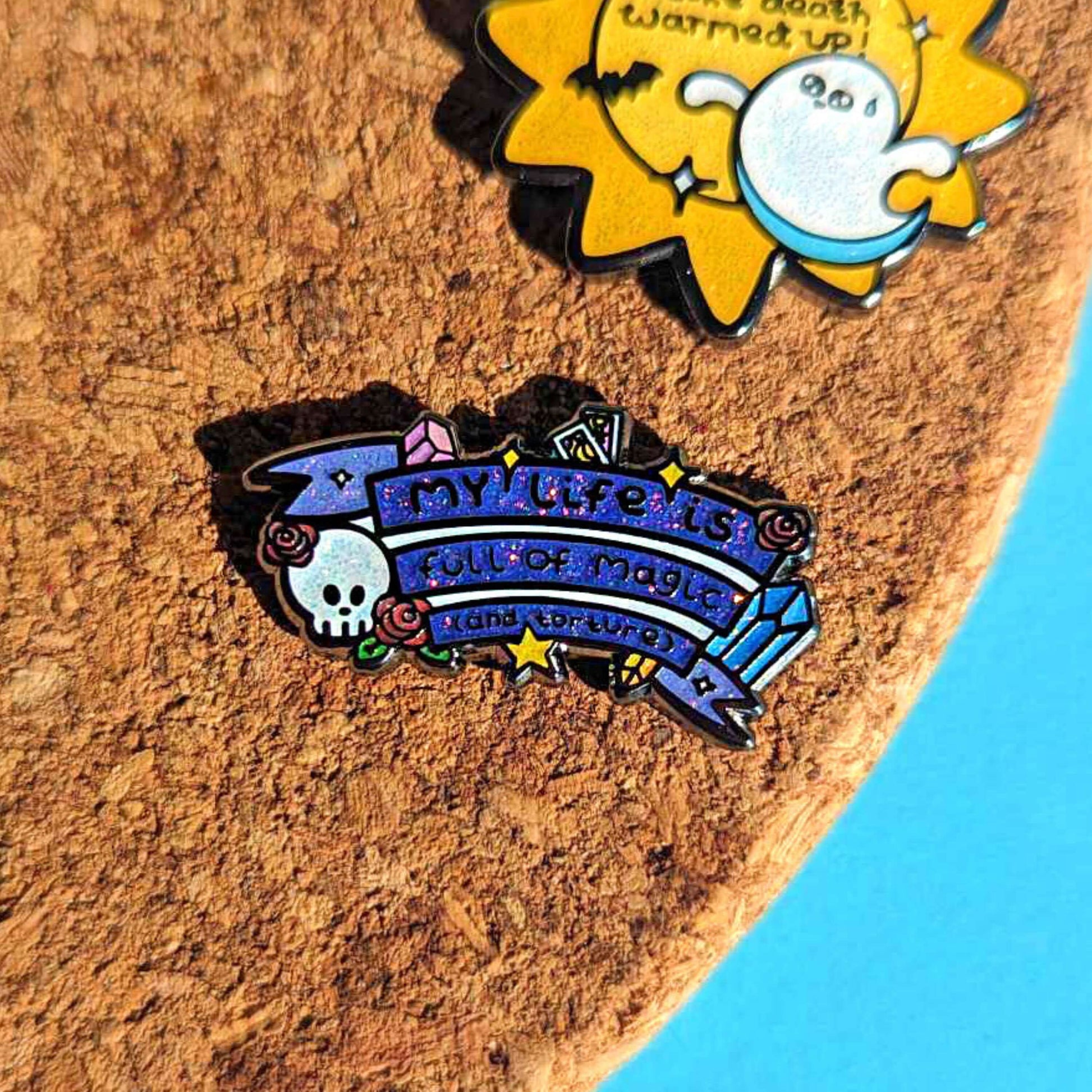 The My Life is Full of Magic & Torture Glitter Enamel Pin pinned onto a cork pin board with other innabox pins. The glittery finish enamel pin badge features a purple banner sash of black text reading 'my life is full of magic (and torture)' with yellow sparkles, crystals, red roses, tarot cards and a skull head.