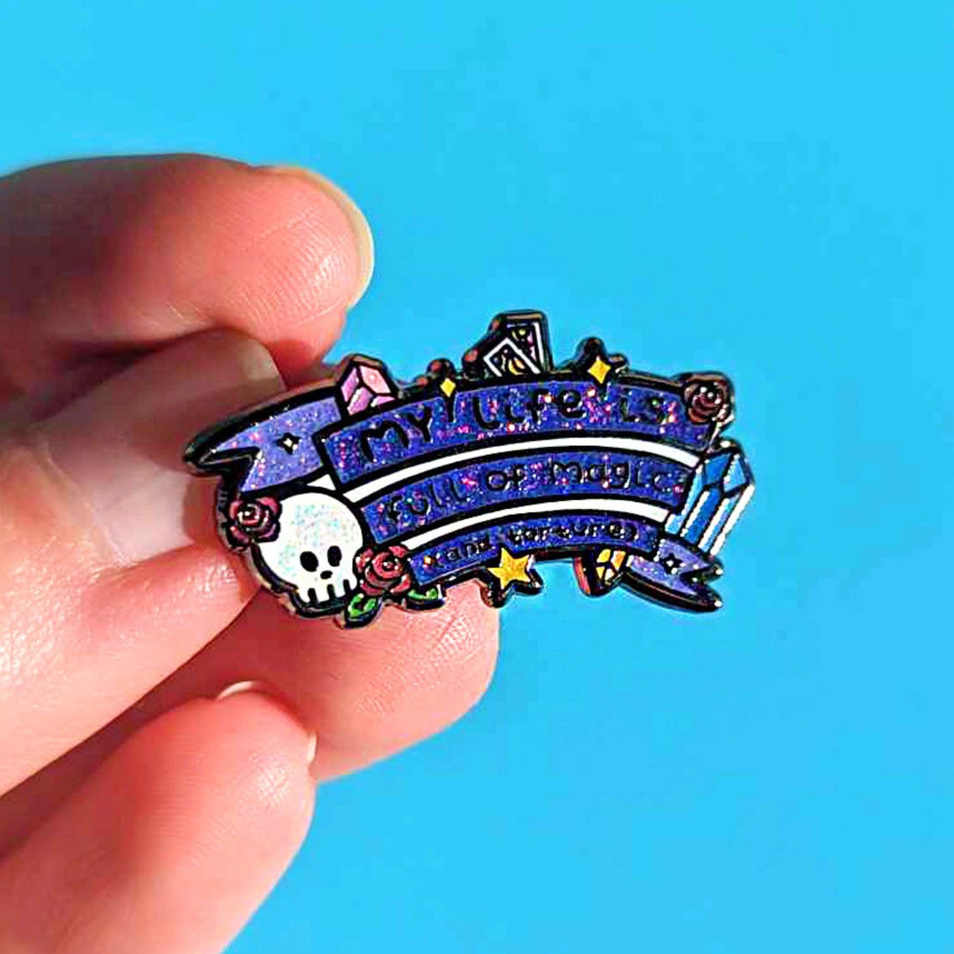 The My Life is Full of Magic & Torture Glitter Enamel Pin held over a blue background. The glittery finish enamel pin badge features a purple banner sash of black text reading 'my life is full of magic (and torture)' with yellow sparkles, crystals, red roses, tarot cards and a skull head.