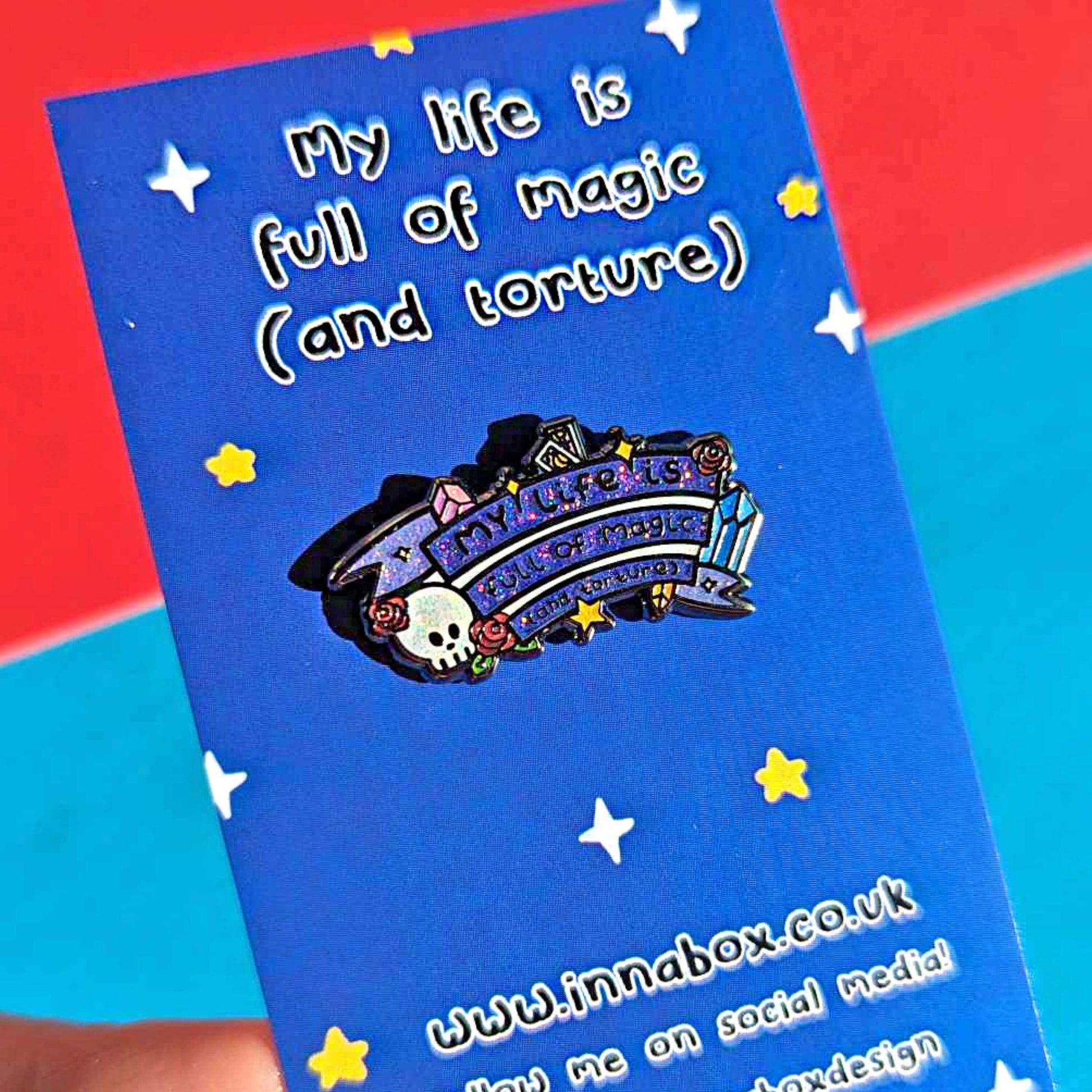 The My Life is Full of Magic & Torture Glitter Enamel Pin on blue starry backing card with the innabox social media handles at the bottom held over a red and blue background. The glittery finish enamel pin badge features a purple banner sash of black text reading 'my life is full of magic (and torture)' with yellow sparkles, crystals, red roses, tarot cards and a skull head.