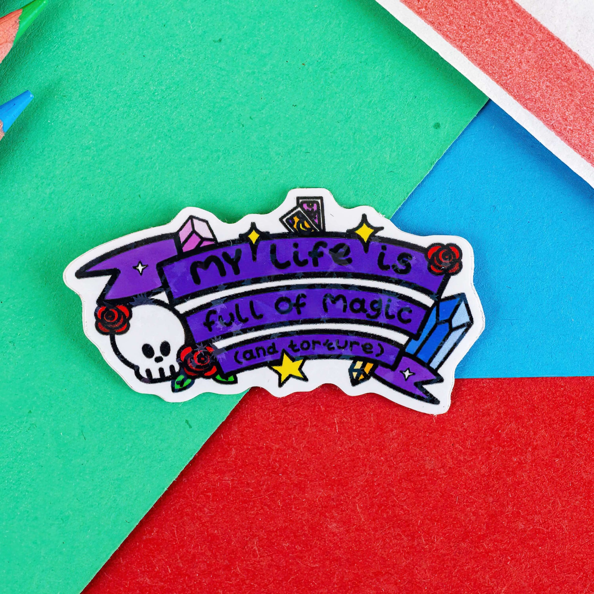 The My Life is Full of Magic & Torture Holographic Sticker on a red, blue and green background with colouring pencils and red stripe candy bag. The holographic stars sticker features a purple banner sash of black text reading 'my life is full of magic (and torture)' with yellow sparkles, crystals, red roses, tarot cards and a skull head.