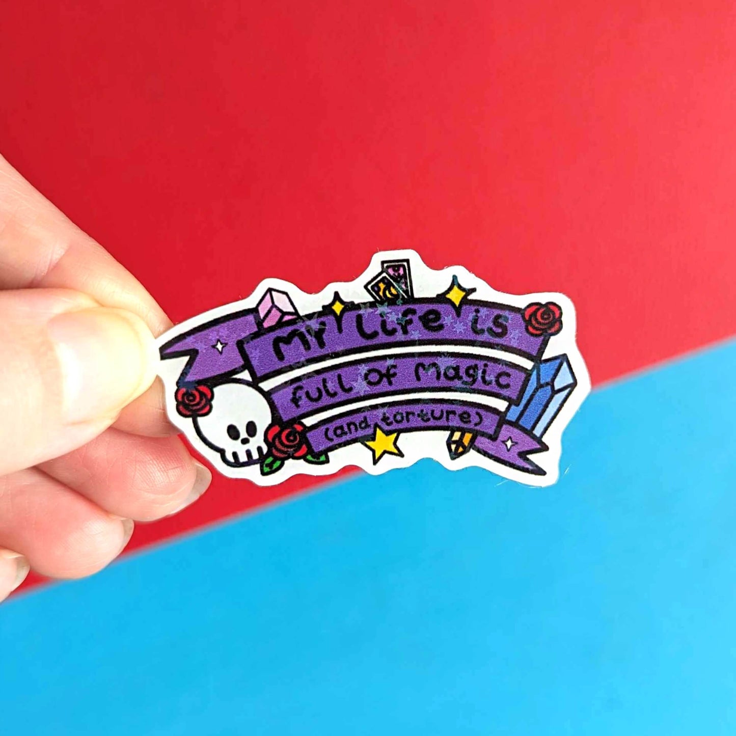 The My Life is Full of Magic & Torture Holographic Sticker held over a red and blue background. The holographic stars sticker features a purple banner sash of black text reading 'my life is full of magic (and torture)' with yellow sparkles, crystals, red roses, tarot cards and a skull head.